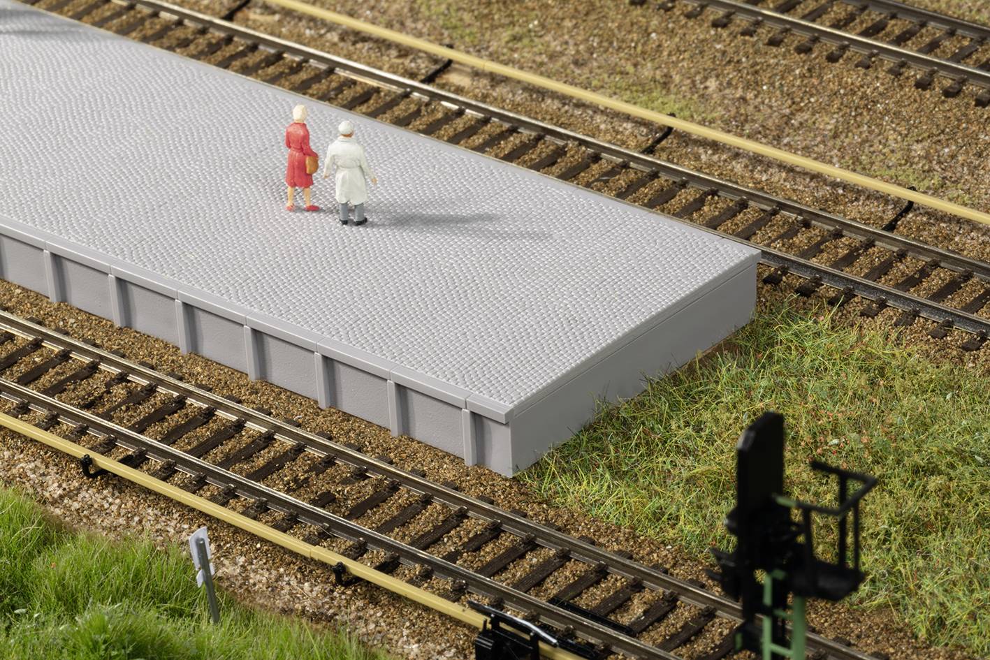 Dve figúry stoja na platforme modelu železnice, obklopené koľajnicami a trávou. Na spodnom okraji obrázka je viditeľný cestný signálny semafor.