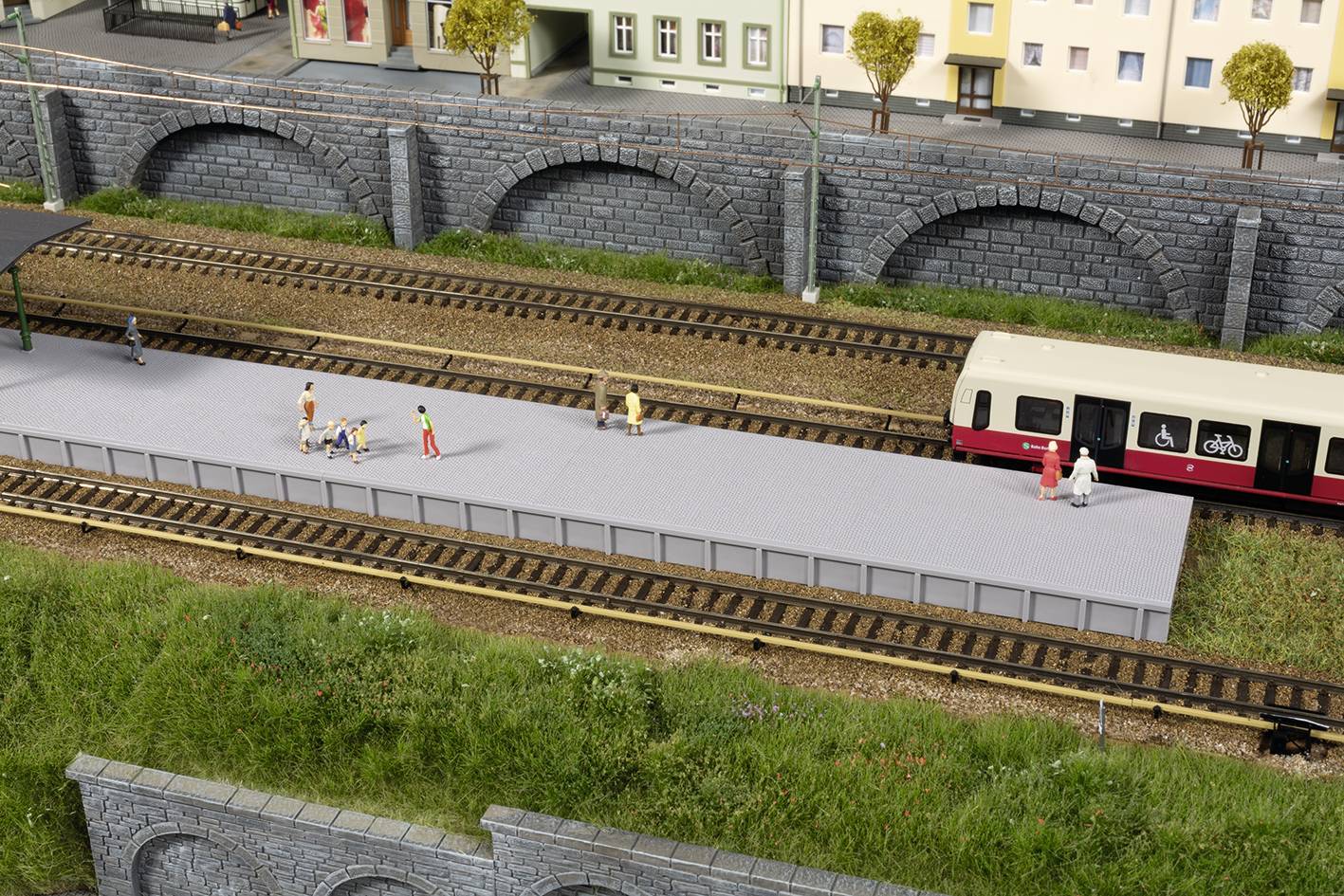 Model železničnej stanice s čakajúcimi figurkami vedľa vlaku. V pozadí je rad domov a kamenný most.