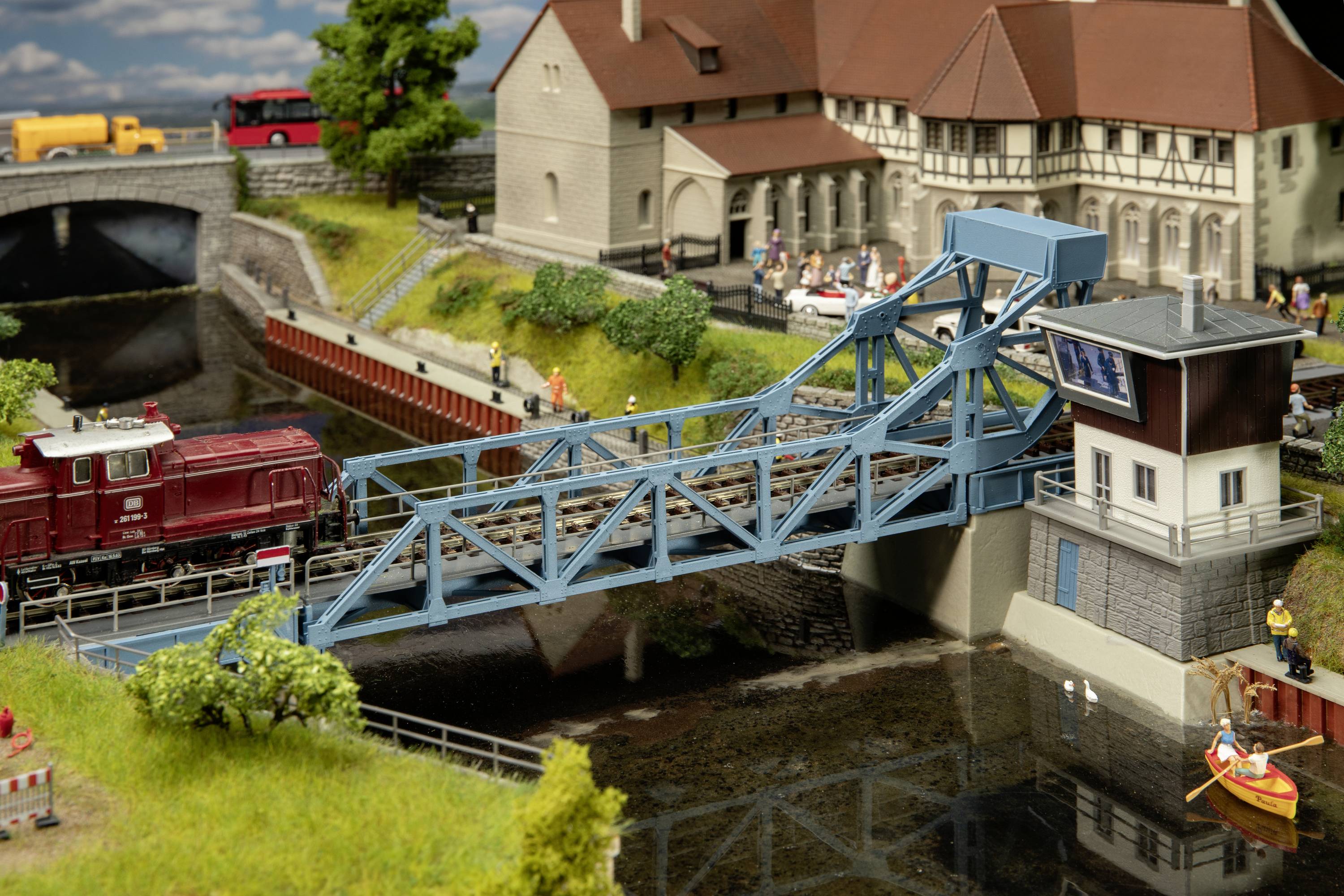Miniatúrna železničná dioráma s pohyblivým mostom, vlakom, detailne spracovanými budovami a krajinou. Scéna pôsobí realisticky a živo.