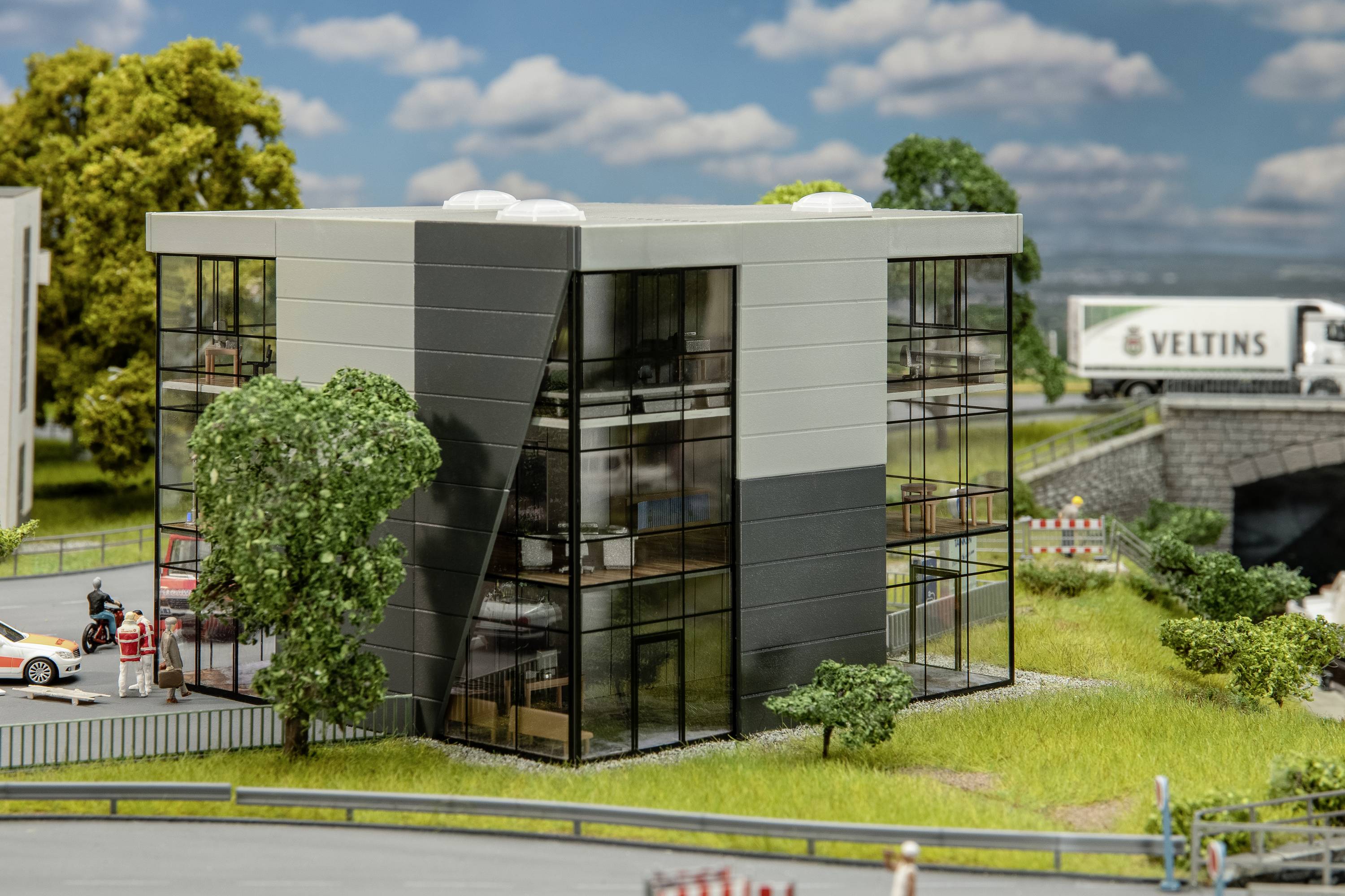Moderná kancelárska budova so sklenenou fasádou v miniatúrnom zobrazení, obklopená zelenými plochami, vedľa cesty a úseku mosta.