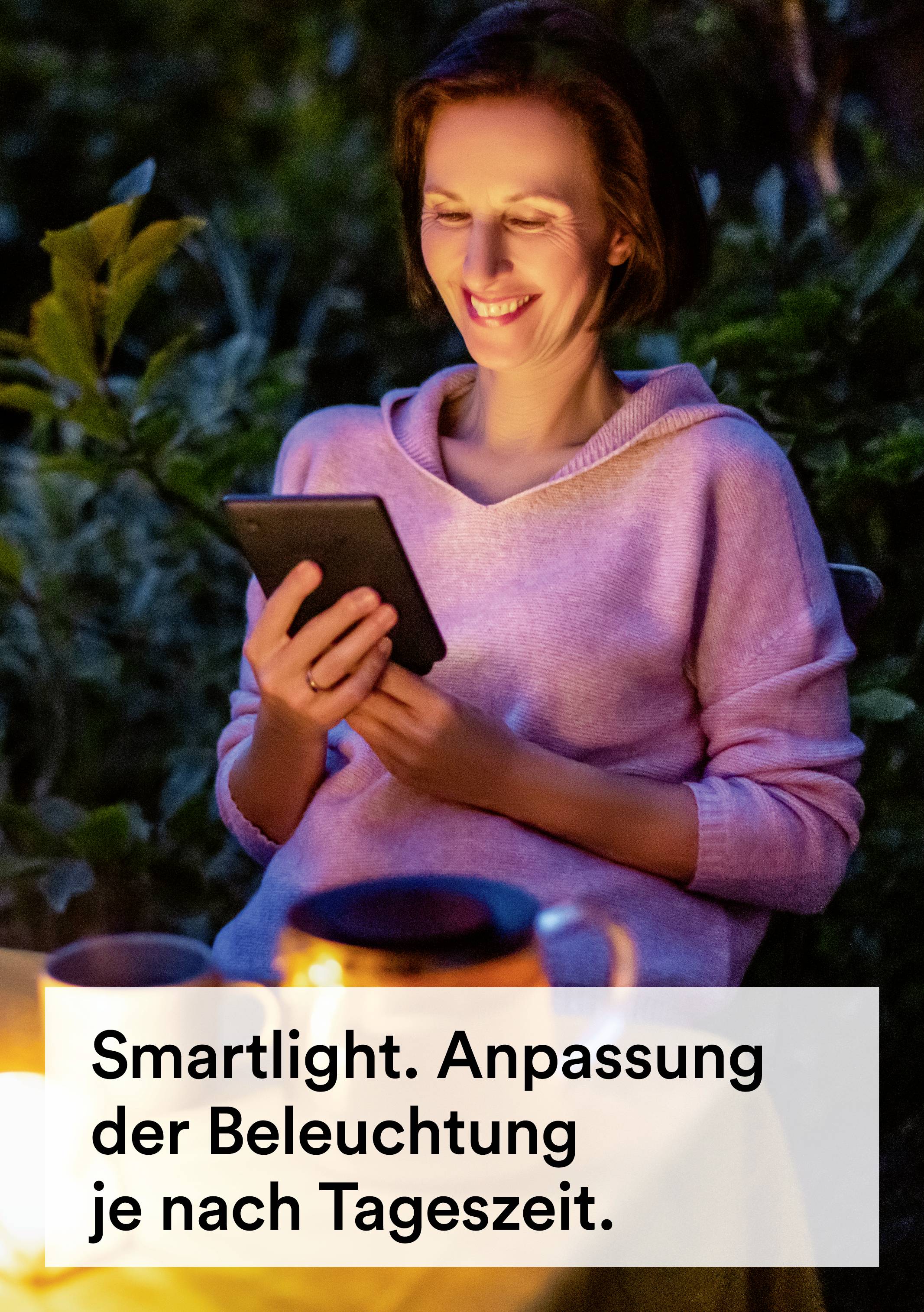 Osoba sedí vonku pri svetle sviečky a číta z tabletu. Text: 'Smartlight. Prispôsobenie osvetlenia podľa dennej doby.'