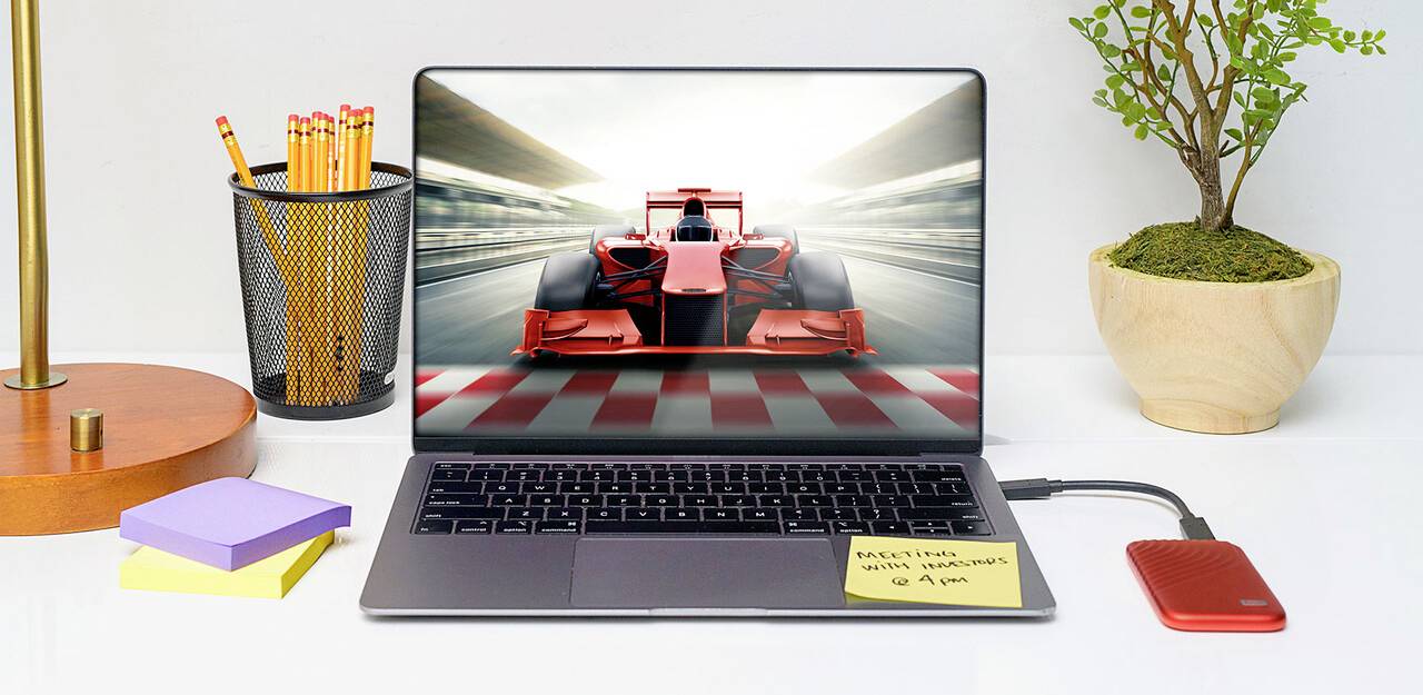 Laptop na pracovnom stole zobrazuje na obrazovke monopost Formuly 1. Vedľa neho sú perá, poznámkový lístok a rastlina.