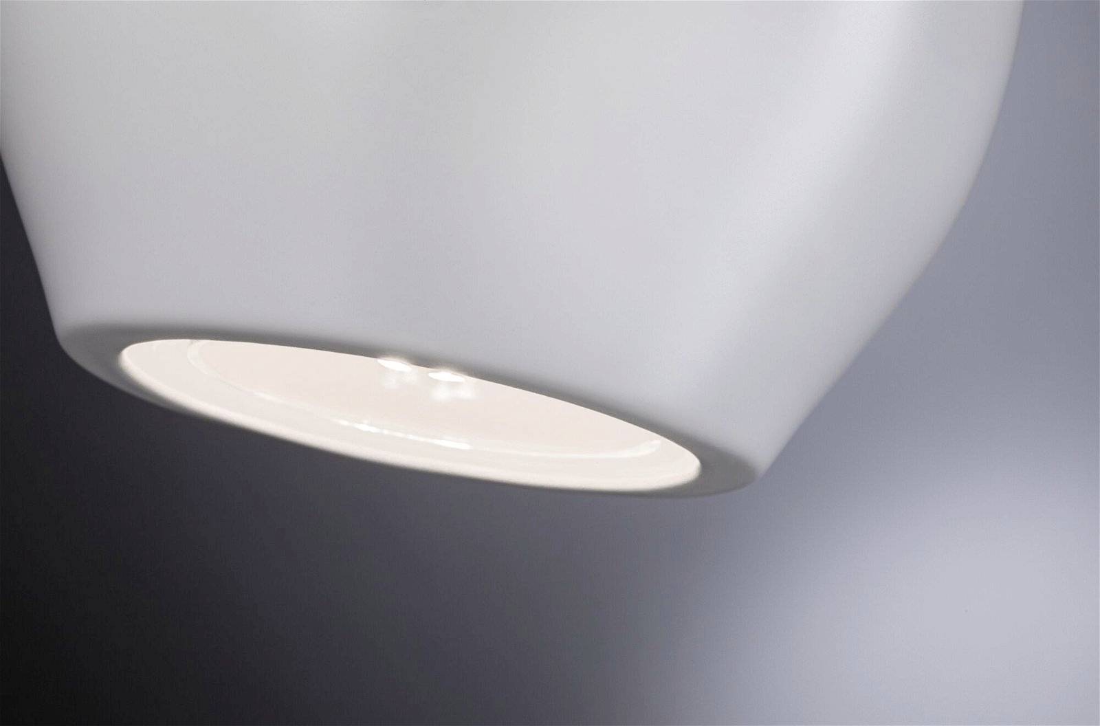 Detailný záber zapnutej modernej lampy s bielym tienidlom. Lampa jemne osvecuje pozadie.