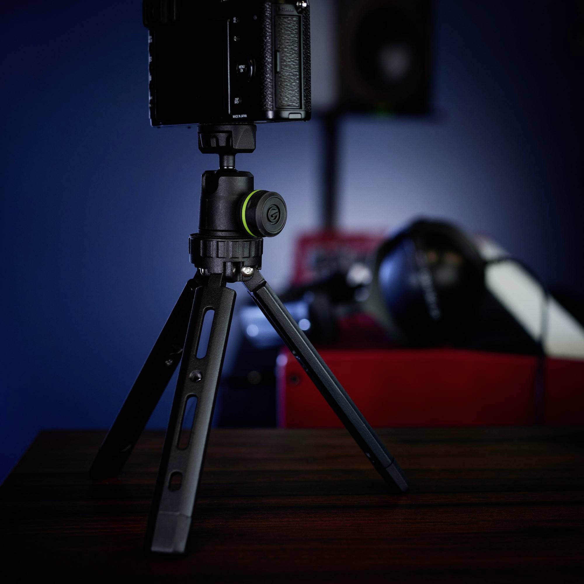 Statív na kameru stojí na stole. V pozadí sú rozmazane viditeľné kamera a ďalšie fotografické príslušenstvo.
