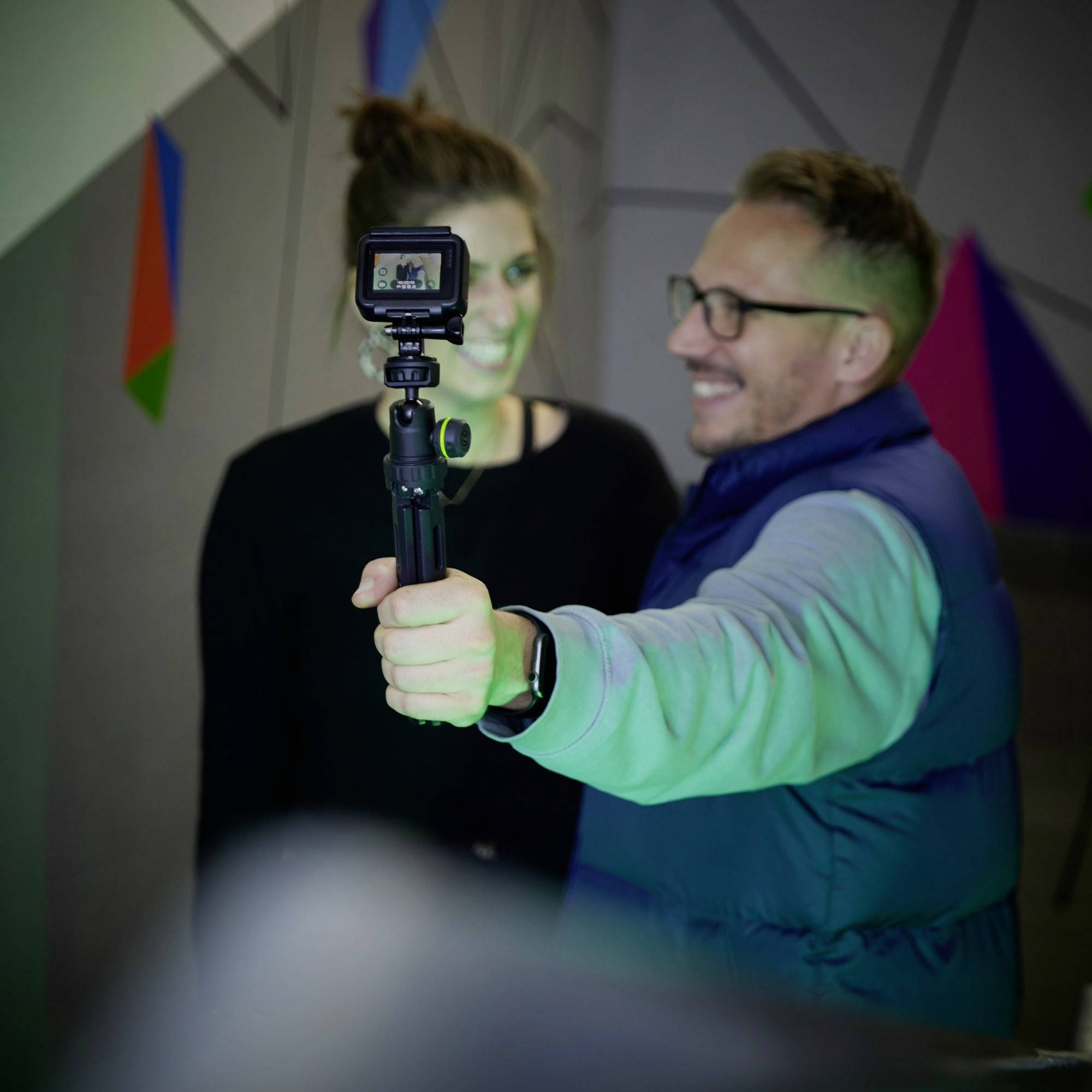 Osoba v popredí drží fotoaparát a usmieva sa, zatiaľ čo si robí selfie s inou osobou v pozadí.