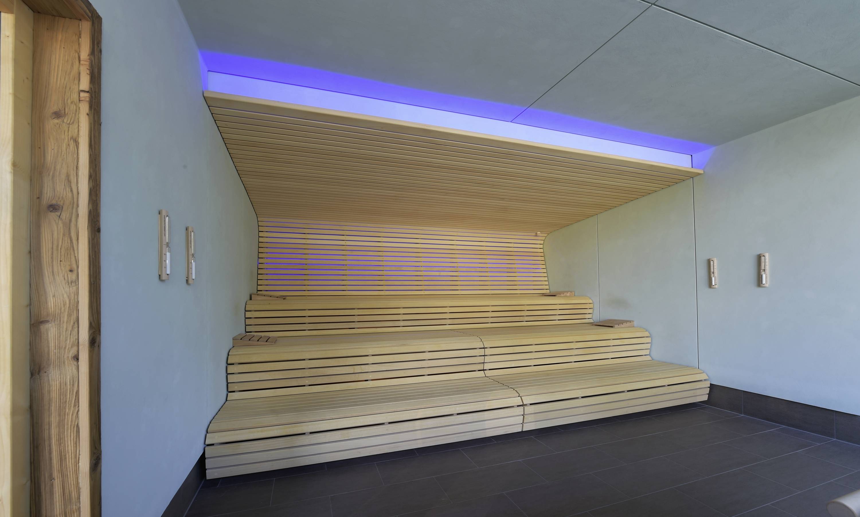 Drevená sauna so stupňovitými sedacími priestormi, modrou LED osvetlením a sivou podlahou.