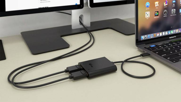 Laptop prepojený s USB hubom, ku ktorému je pripojených niekoľko káblov. V pozadí sú dva monitory zobrazujúce plochy aplikácií.