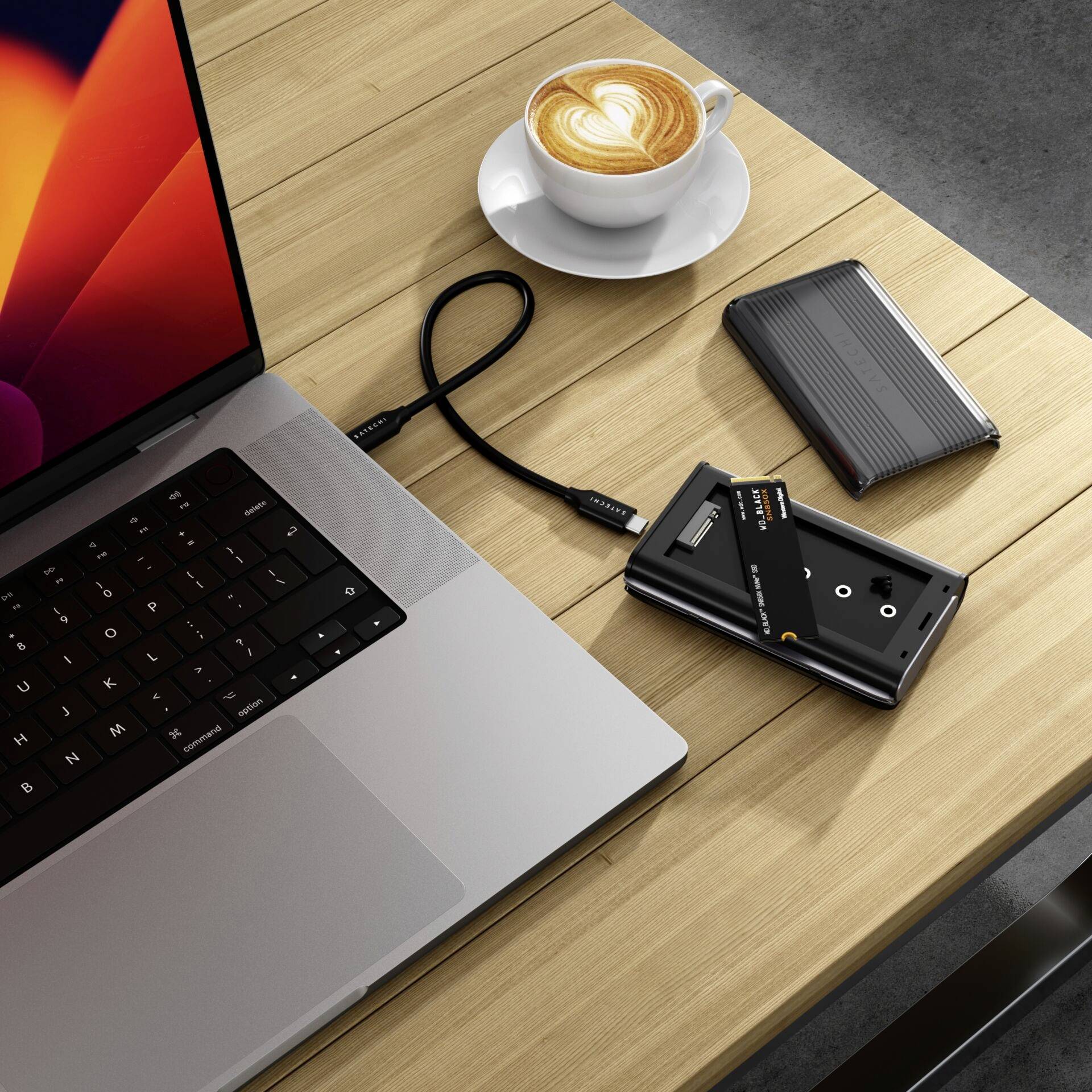 Na drevenom stole stoja laptop, šálka cappuccina, externý disk, čítačka kariet a USB kábel.