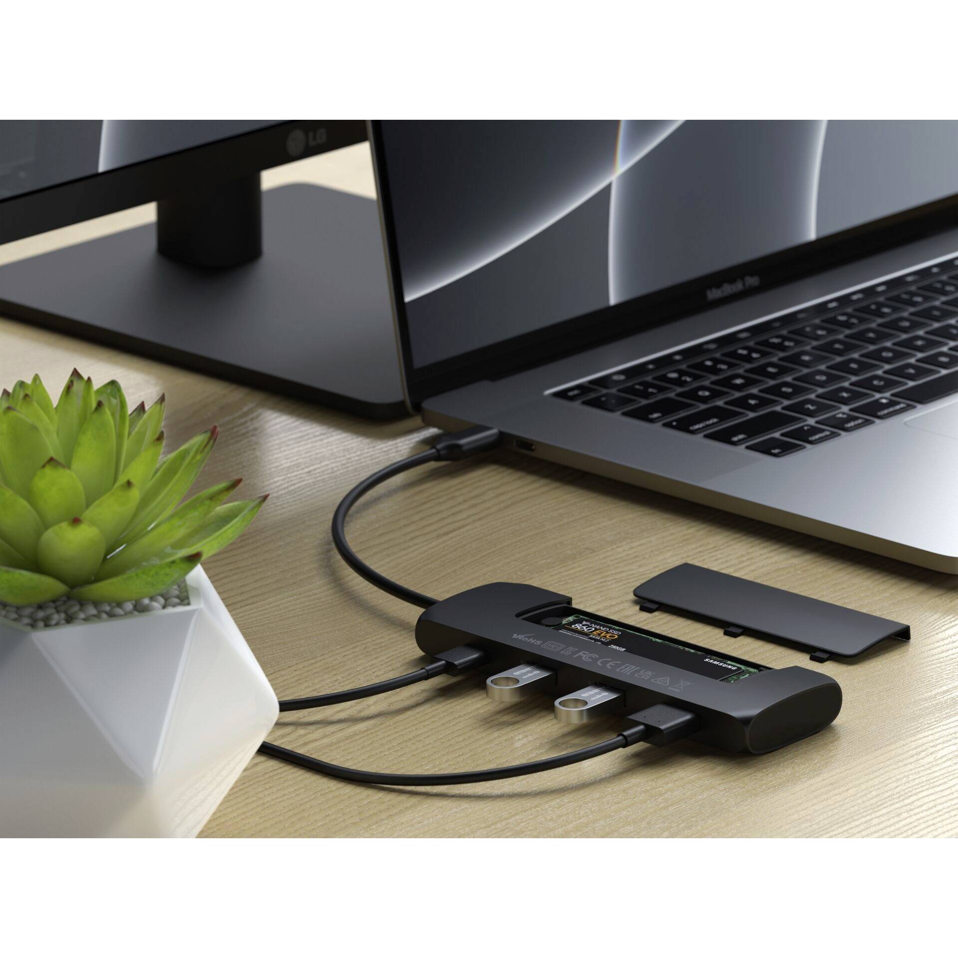 Laptop je pripojený k USB rozbočovaču, ku ktorému sú pripojené káble a USB disky. Vedľa stojí monitor a rastlina.