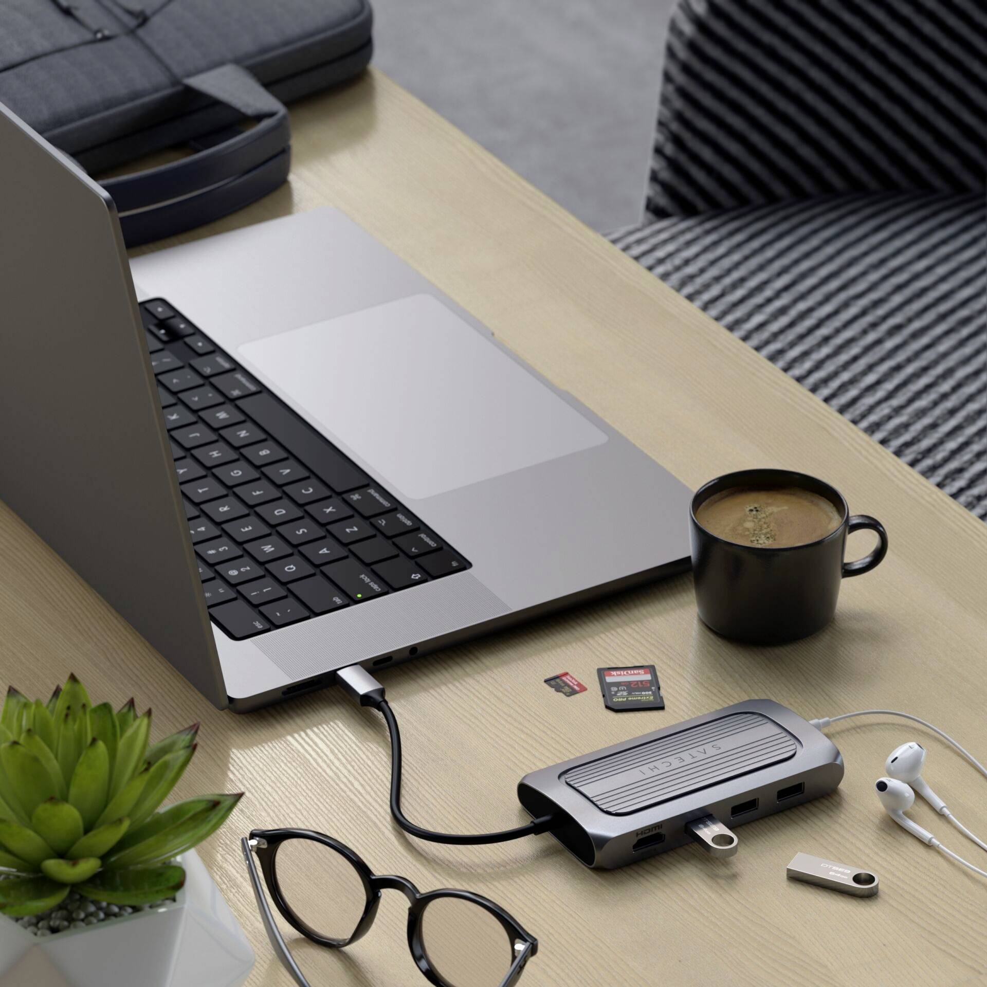 Laptop na stole, pripojený k USB rozbočovaču, vedľa šálka kávy, pamäťová karta fotoaparátu, okuliare a rastlina.