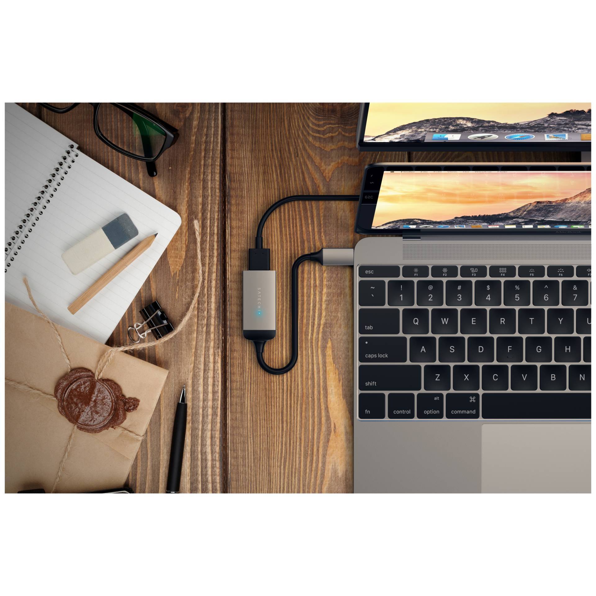 Laptop prepojený s USB zariadením, na pracovnom stole s poznámkovým blokom, okuliarmi a obálkou. Druhý monitor zobrazuje výhľad na more.