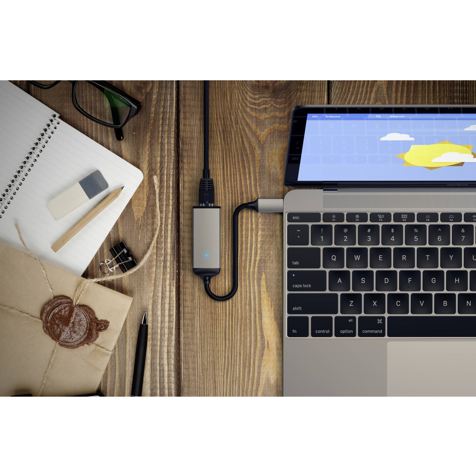 Laptop na drevenej doske stola, pripojený k USB rozbočovaču; vedľa notes, obálka s voskovou pečaťou, okuliare a guma.