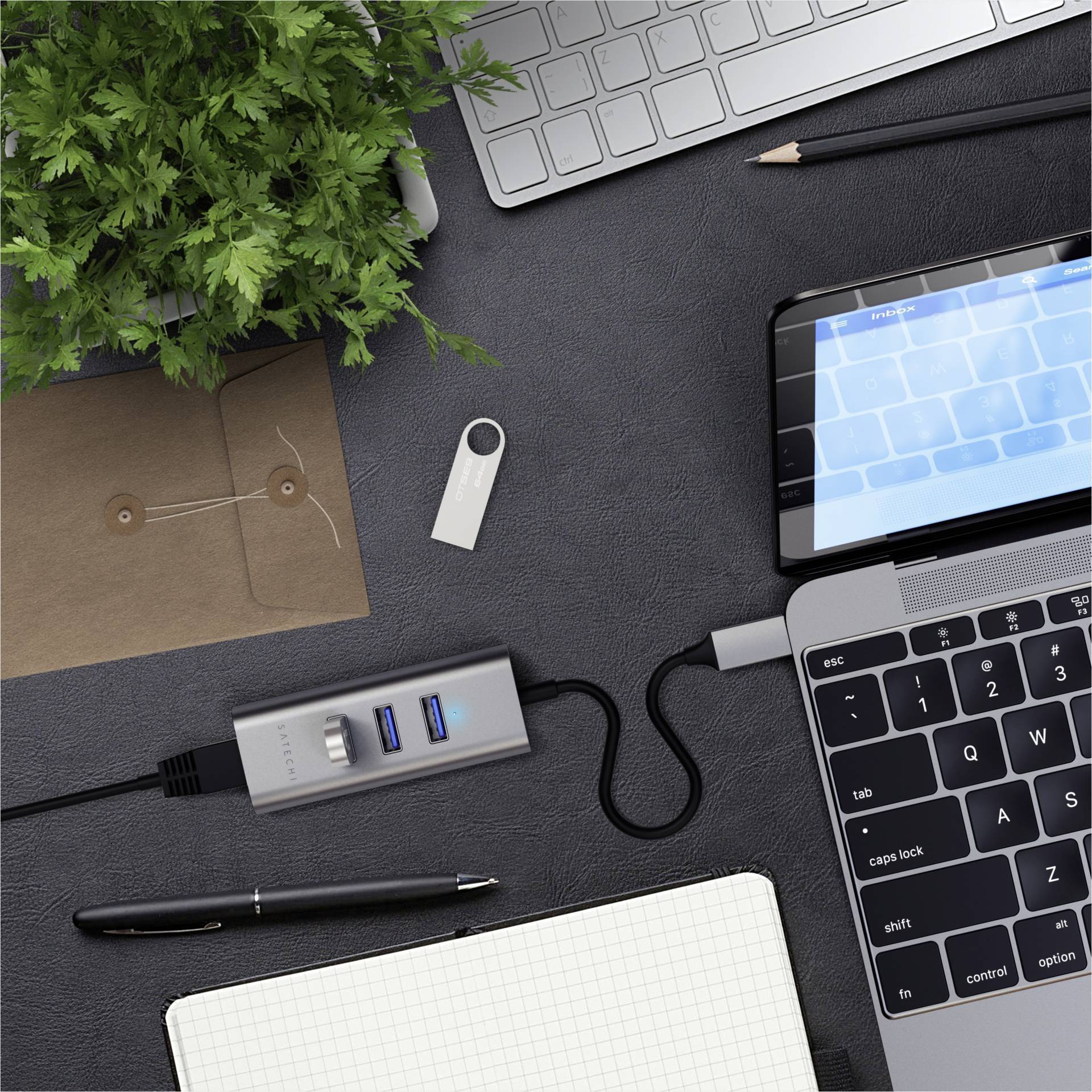Pracovisko s laptopom a pripojeným USB hubom, perom, rastlinami a listami; scéna technologického pracoviska.