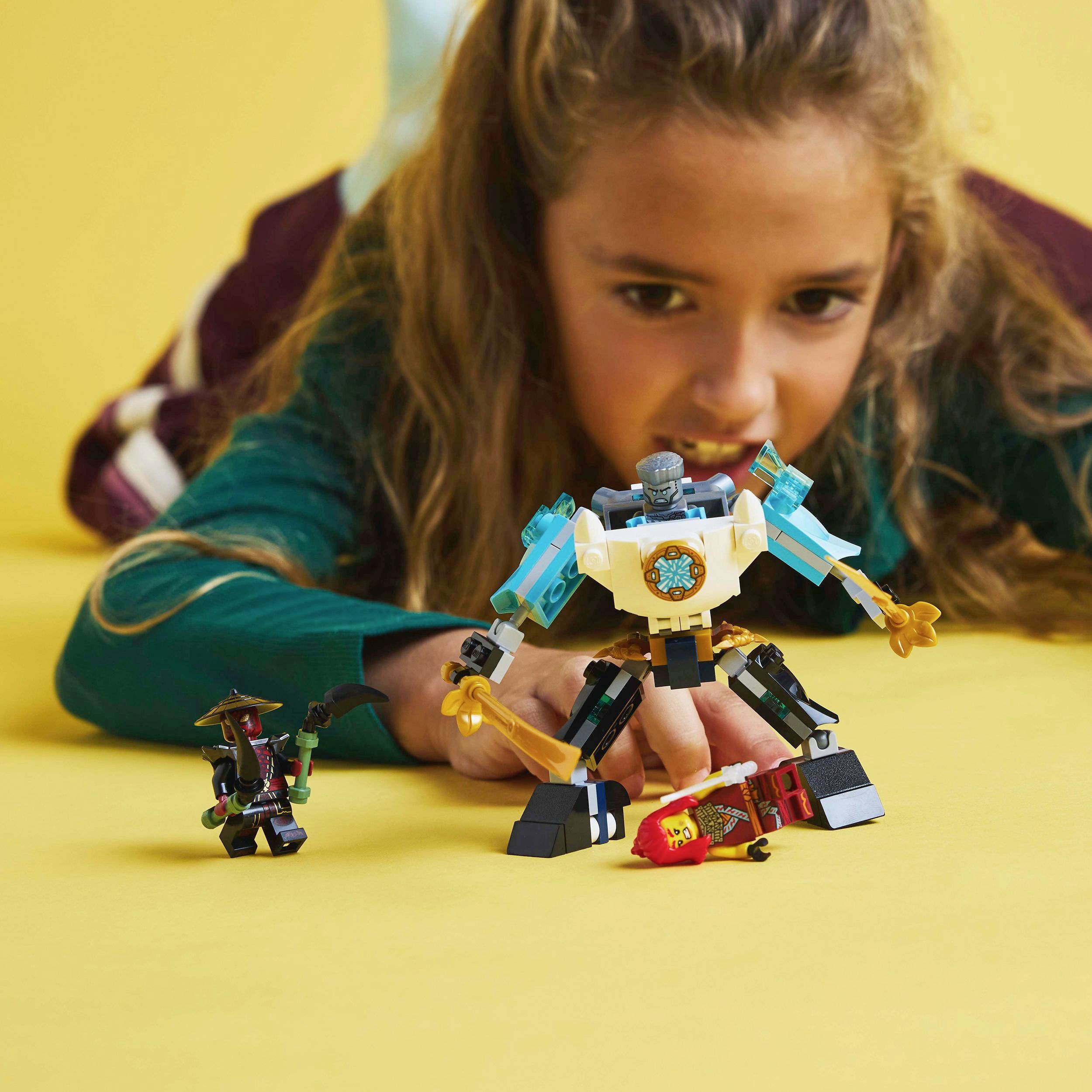 Dieťa sa sústredeným výrazom hrá s farebnými lego robotmi na žltom stole, ktoré majú rôzne tvary a doplnky.