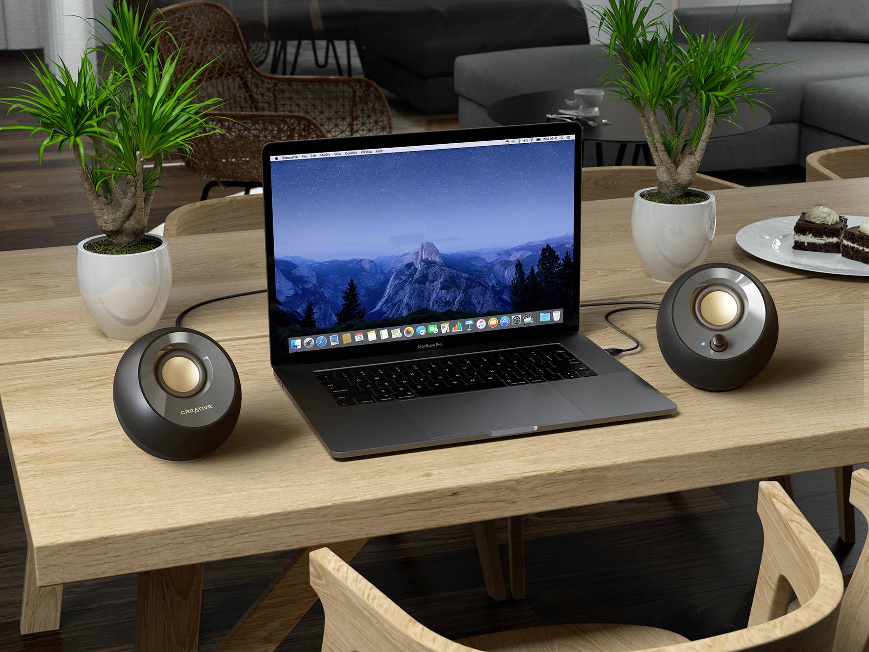 Laptop na drevenej doske zobrazuje pozadie s horskou krajinou. Vedľa neho stoja dva reproduktory a tri izbové rastliny.