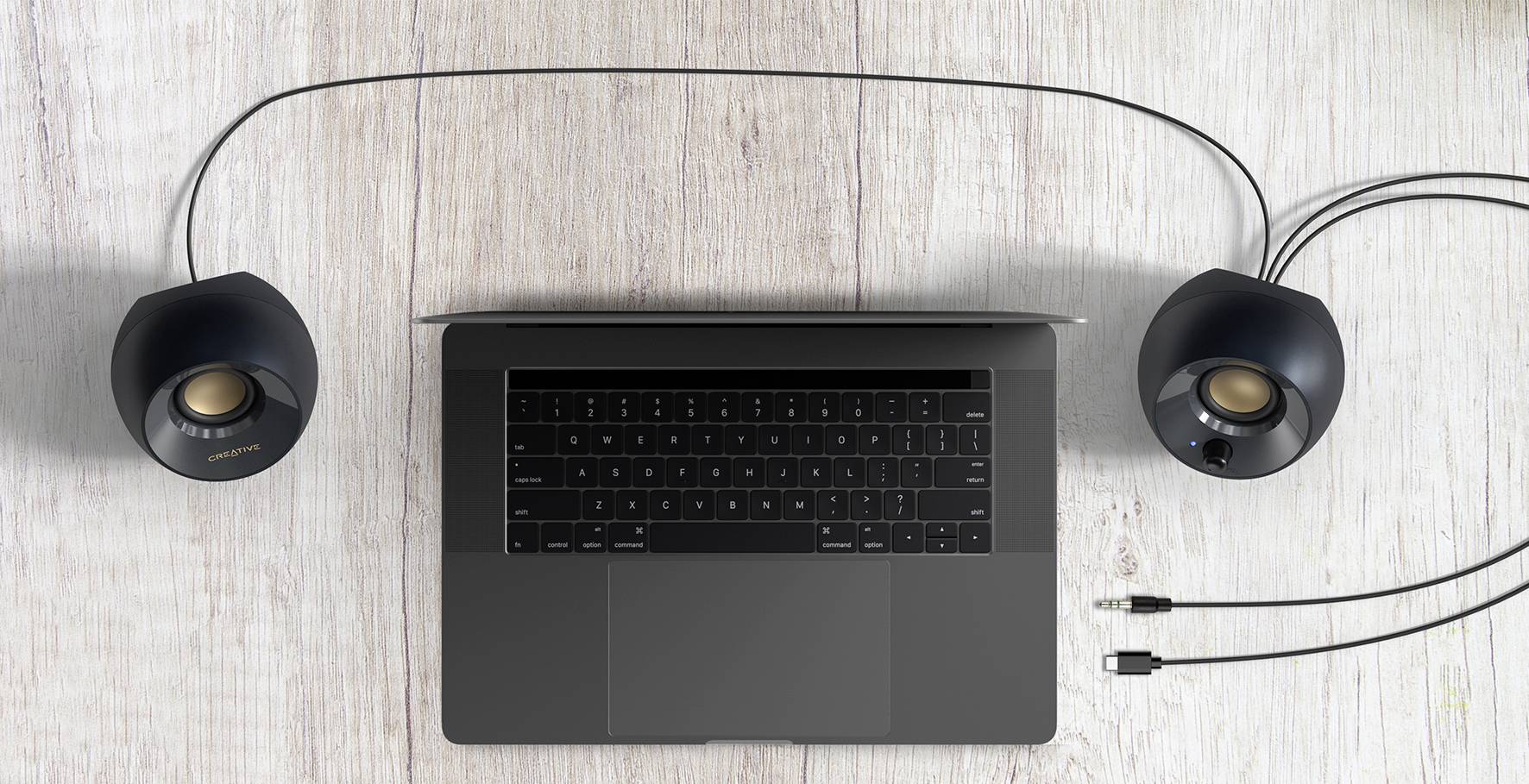 Laptop s čiernou klávesnicou na svetlom drevenom stole, po stranách obklopený dvoma čiernymi reproduktormi, ktoré sú prepojené káblami.