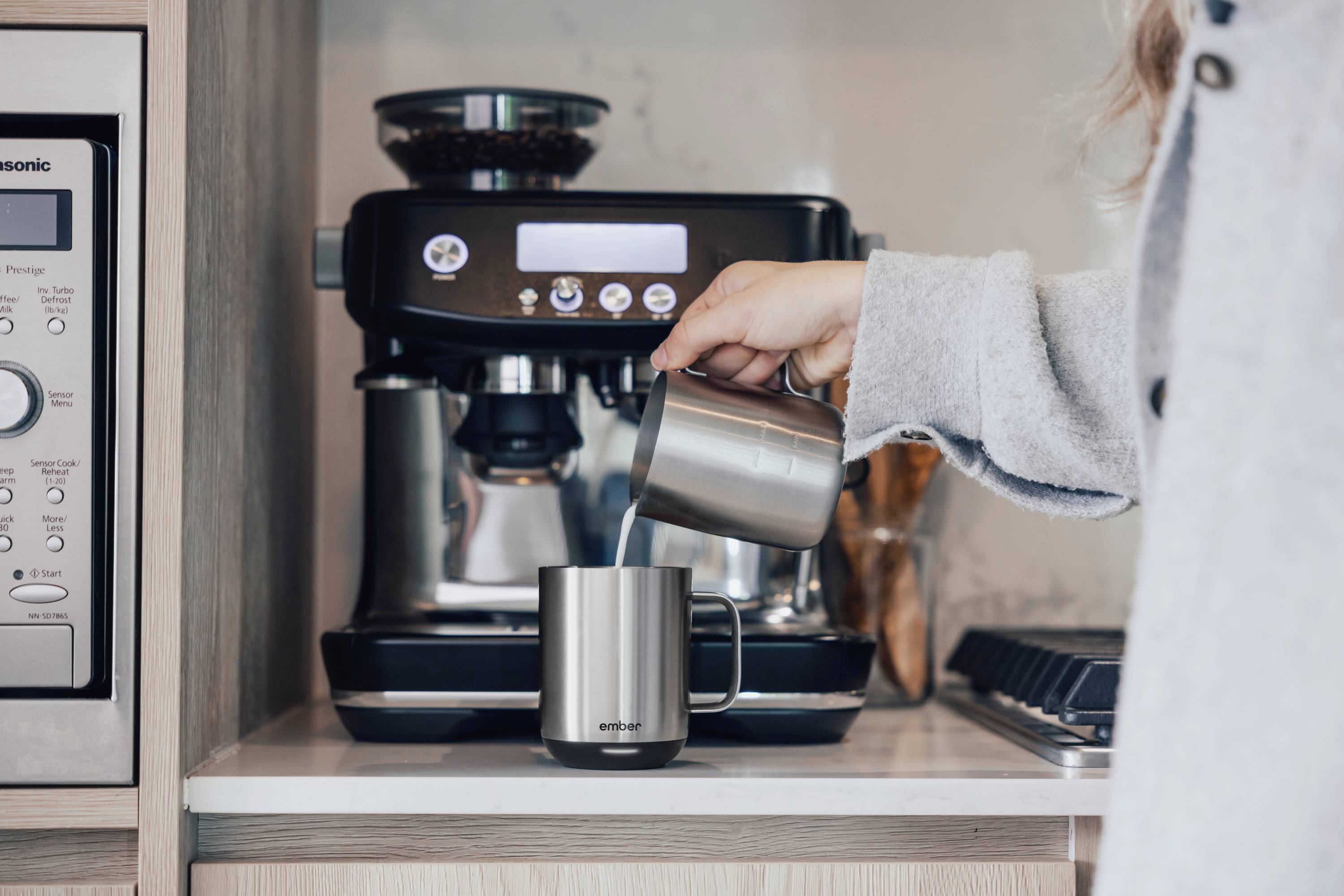 Osoba nalieava mlieko do šálky pred espresso strojom v kuchyni. V pozadí sú viditeľné kuchynské spotrebiče.