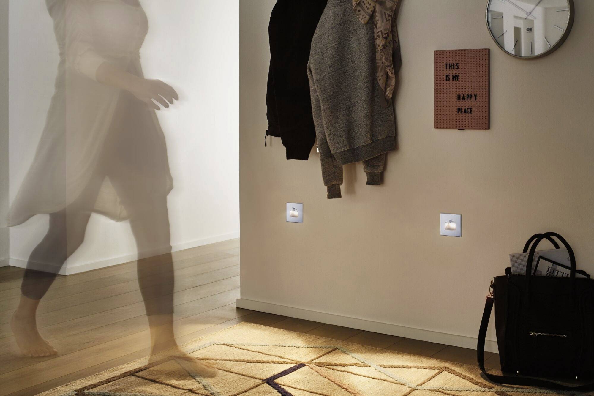 Nejasná postava prechádza okolo steny, na ktorej visia dve bundy. Na stene je vidieť tabuľku s nápisom "This is my happy place".