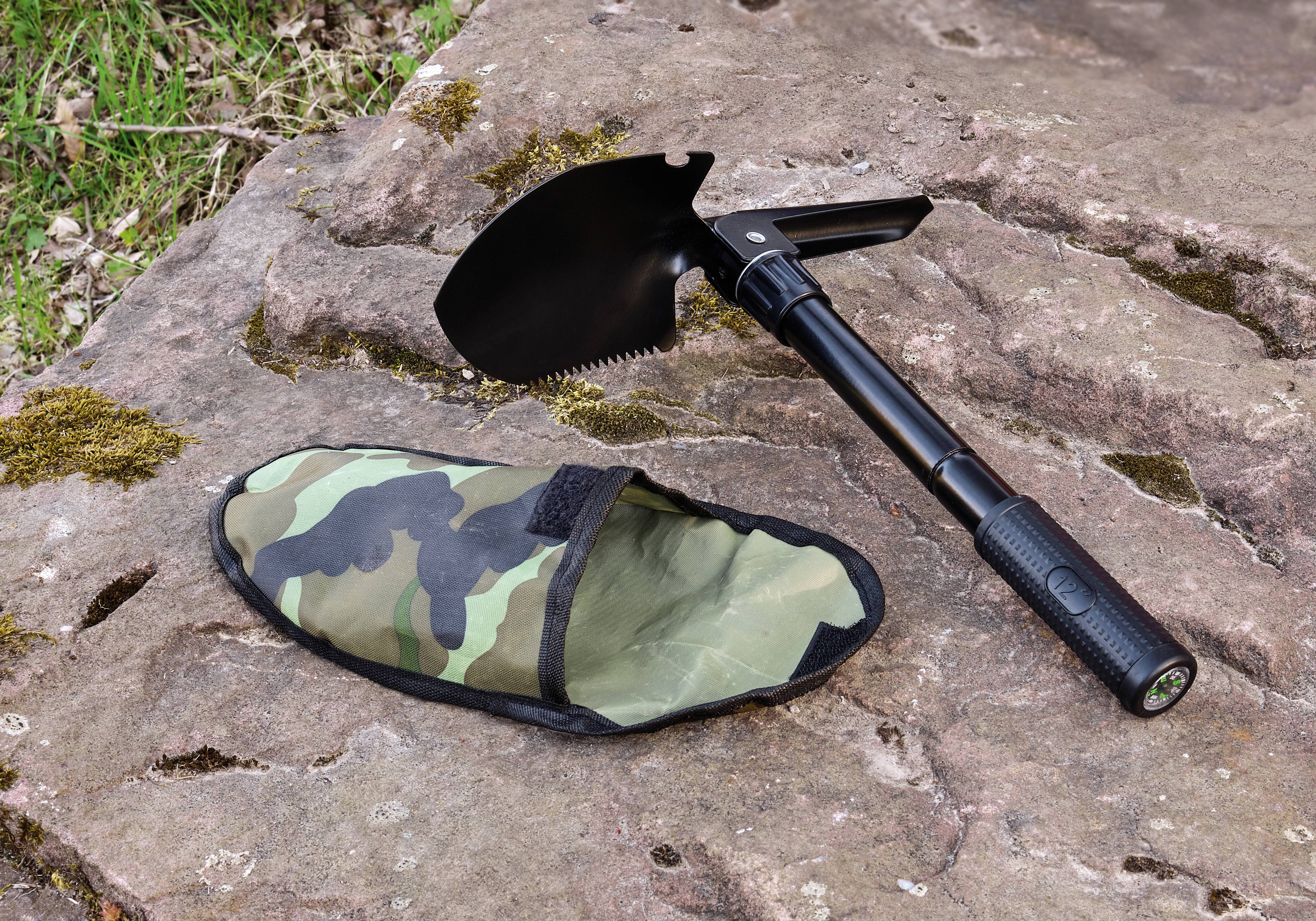 Skladacia lopata so zúbkatým okrajom a čiernou rukoväťou, položená na skalách. Maskáčový obal vedľa nej. Ideálna pre outdoorové použitie.