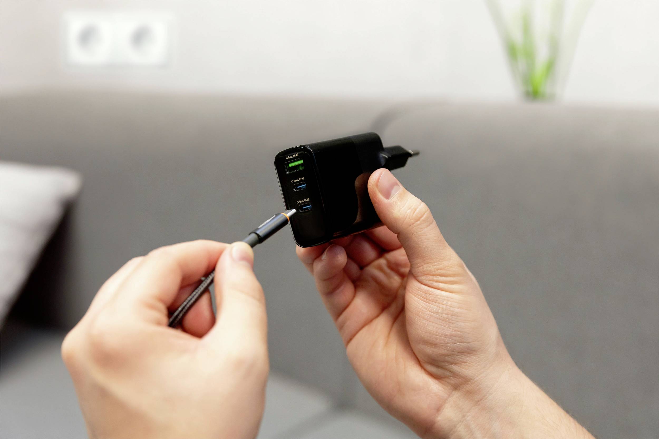 Osoba drží multifunkčnú nabíjačku s viacerými USB portami v ruke a zapája do nej kábel. V pozadí je vidieť pohovku.