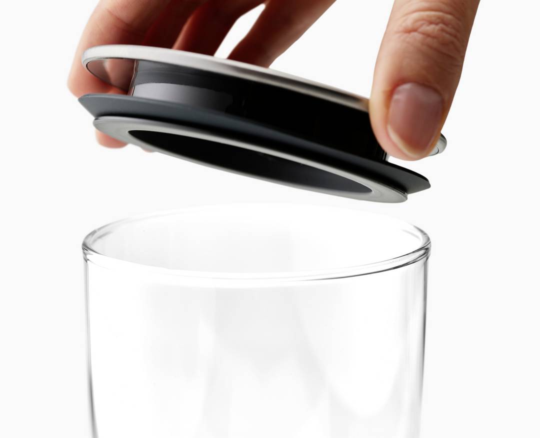 Ruka dvíha kruhové veko zo sklenice, ktorá stojí na bielom pozadí.