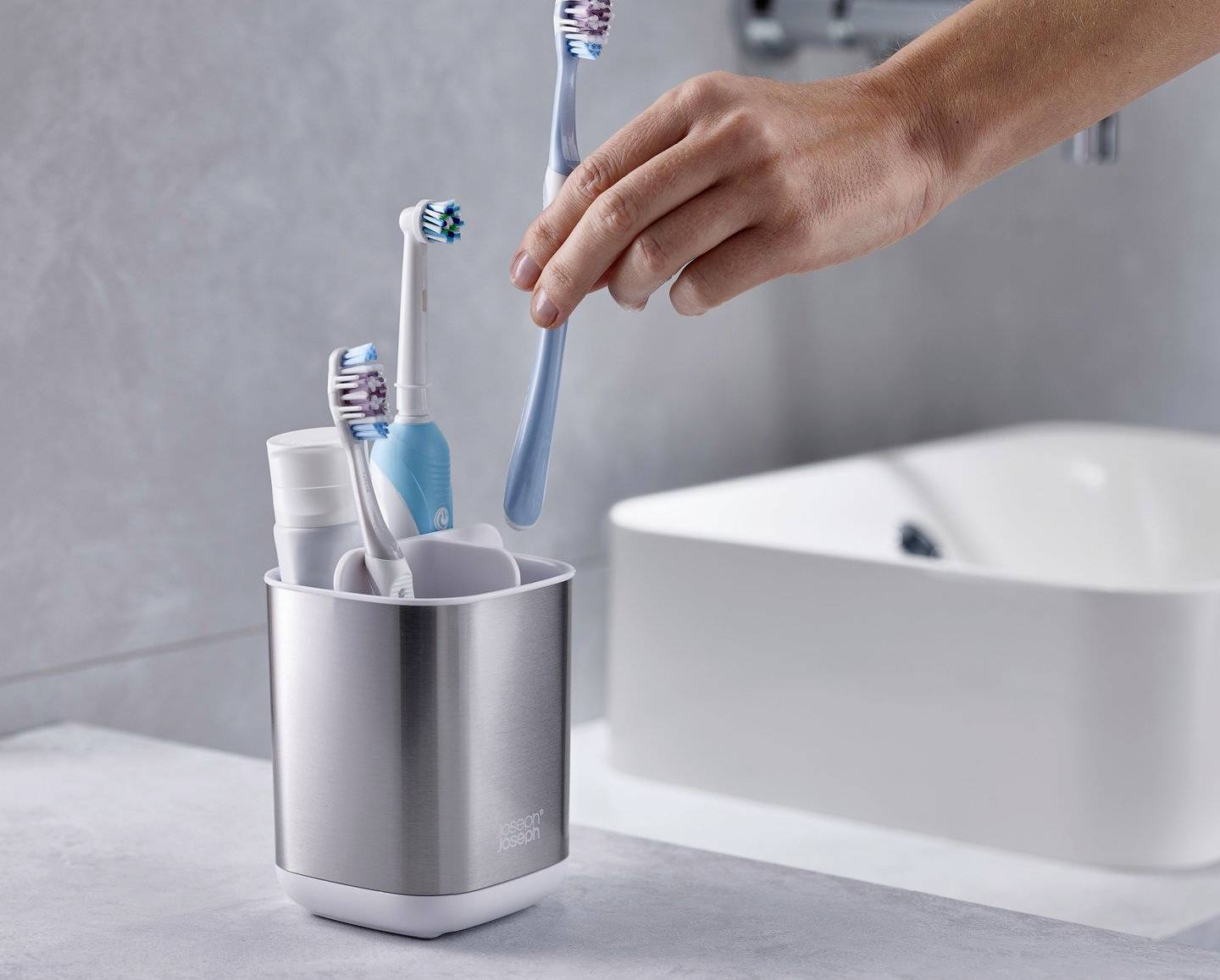 Ruka vyberá modrú zubnú kefku z nerezového pohára, ktorý obsahuje aj ďalšie zubné kefky a zubnú pastu. V pozadí je vidieť umývadlo.