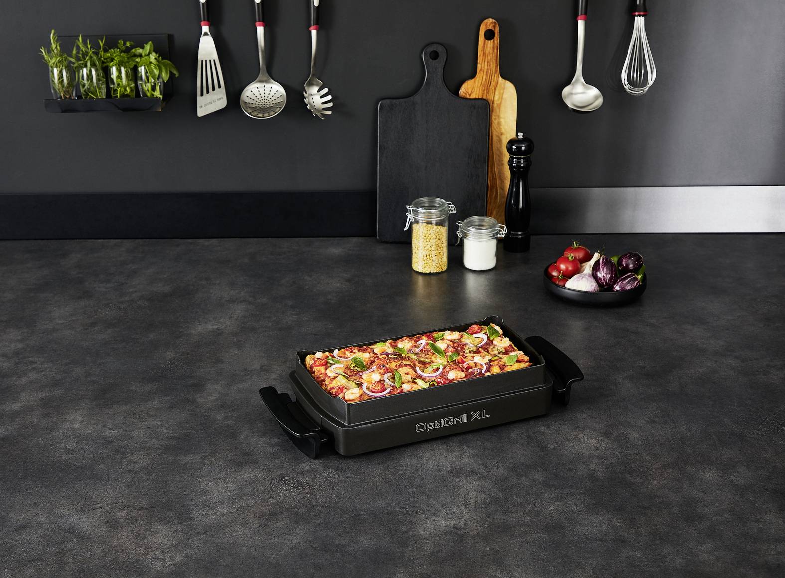 Veľká plnená grilovacia misa s čerstvou zeleninou a bylinkami na kuchynskej pracovnej doske. V pozadí visia kuchynské nástroje.