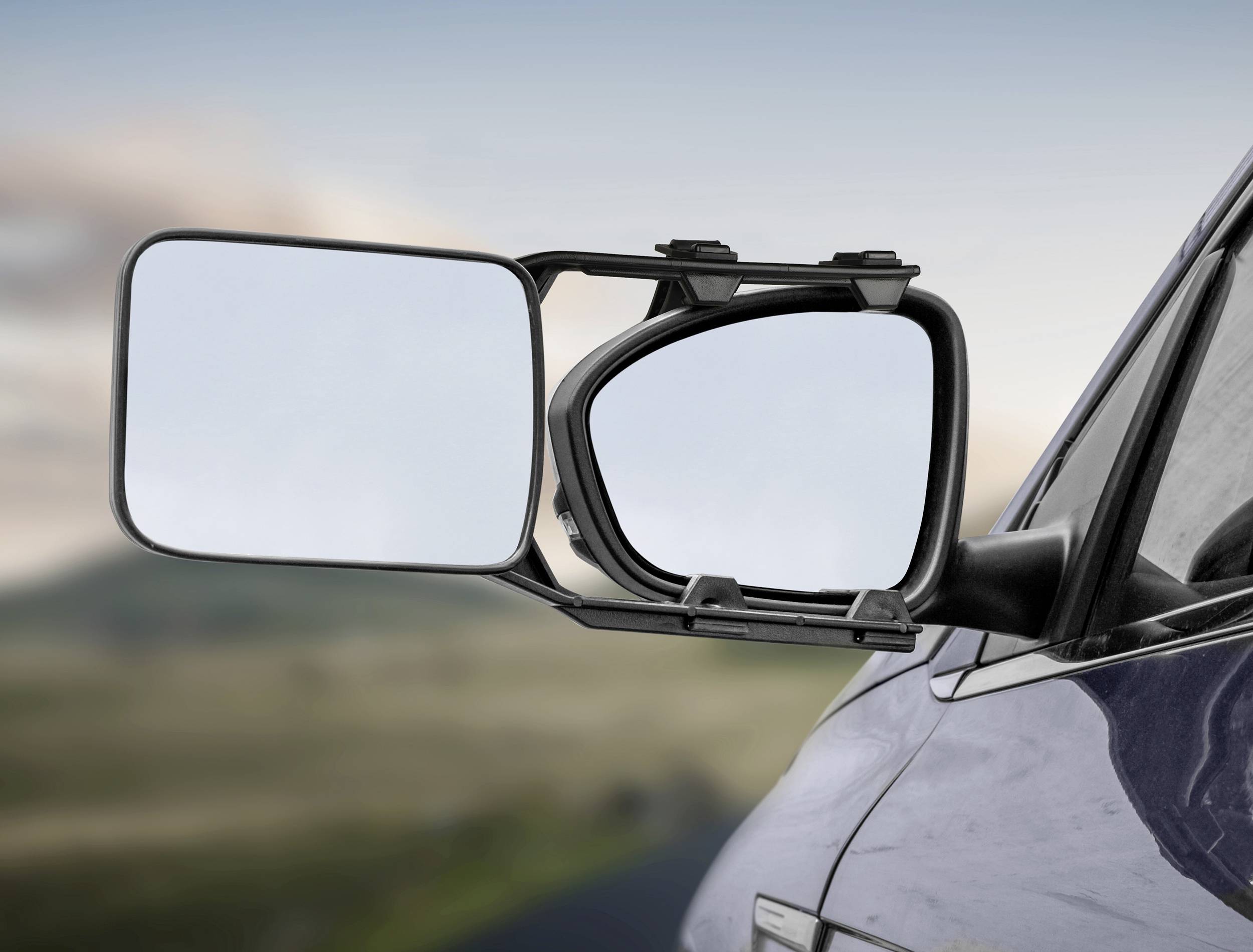 Bočný pohľad na auto s dodatočným zrkadlom pre príves, ktoré kombinuje dva zrkadlá s cieľom rozšíriť zorné pole vodiča. V pozadí je rozmazaná krajina.