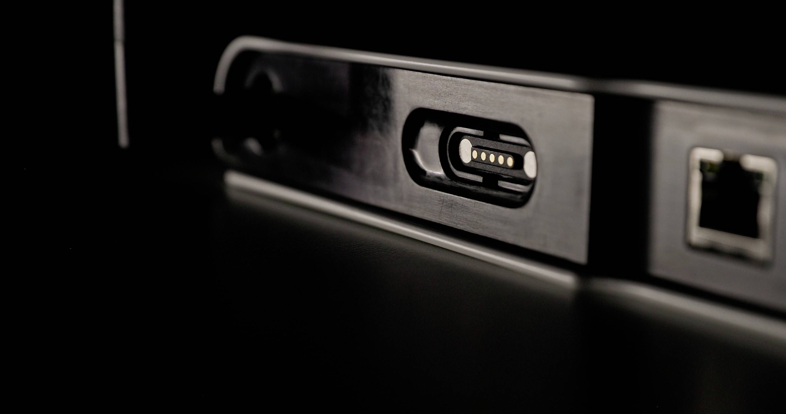 Detailný záber magnetického napájacieho konektora na zariadení v tmavom prostredí, vedľa ktorého je viditeľný ethernetový port.