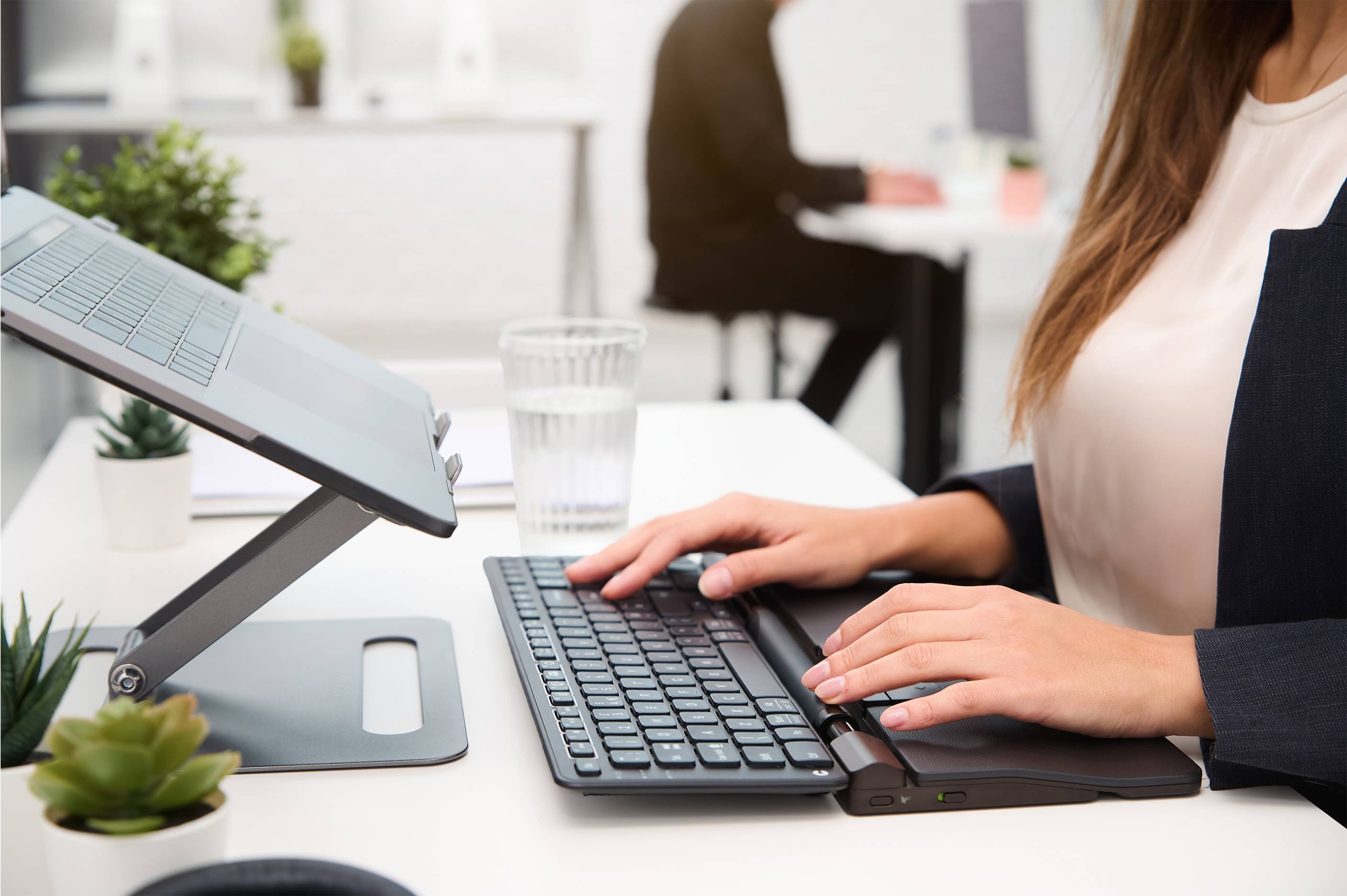 Jedna osoba pracuje pri stole s podstavcom na laptop a klávesnicou; v pozadí sedí ďalšia osoba pri podobnom pracovisku.