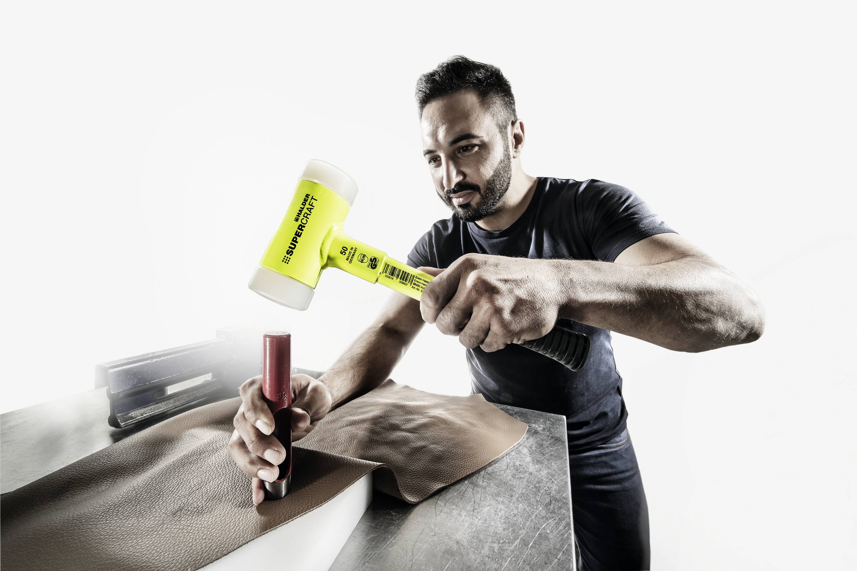 Muž opracováva kus kože žltým kladivom na pracovnom stole. Sústreďuje sa na svoju manuálnu prácu.