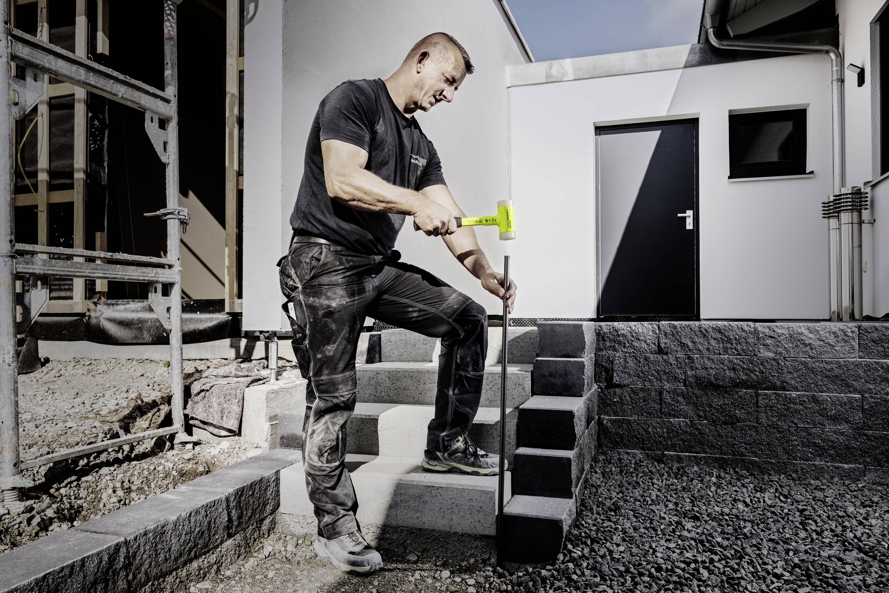 Muž v pracovnom oblečení kladie kladivom malé dlažobné kocky na stavenisku pri dokončovaní schodiska.