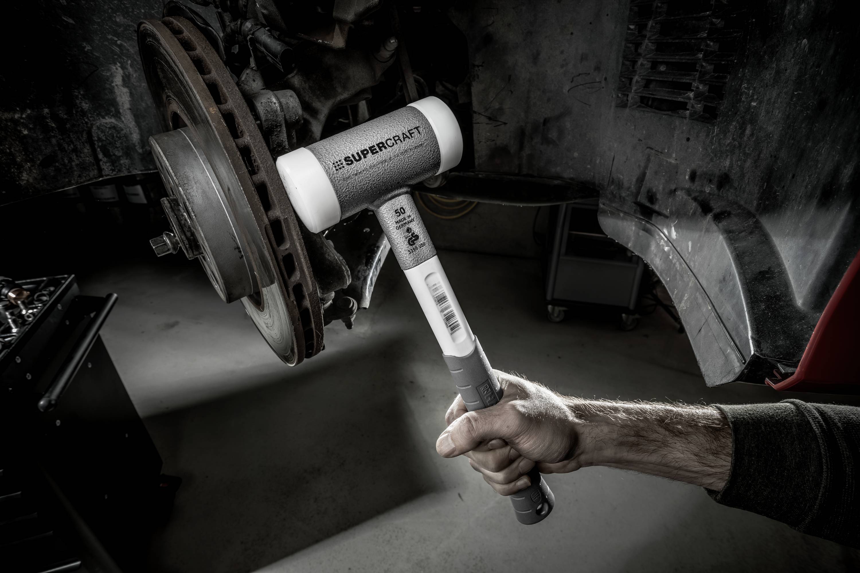 Jedna ruka drží biely kladivá, ktorý je namierený na brzdový kotúč. V pozadí sú viditeľné automobilové diely.