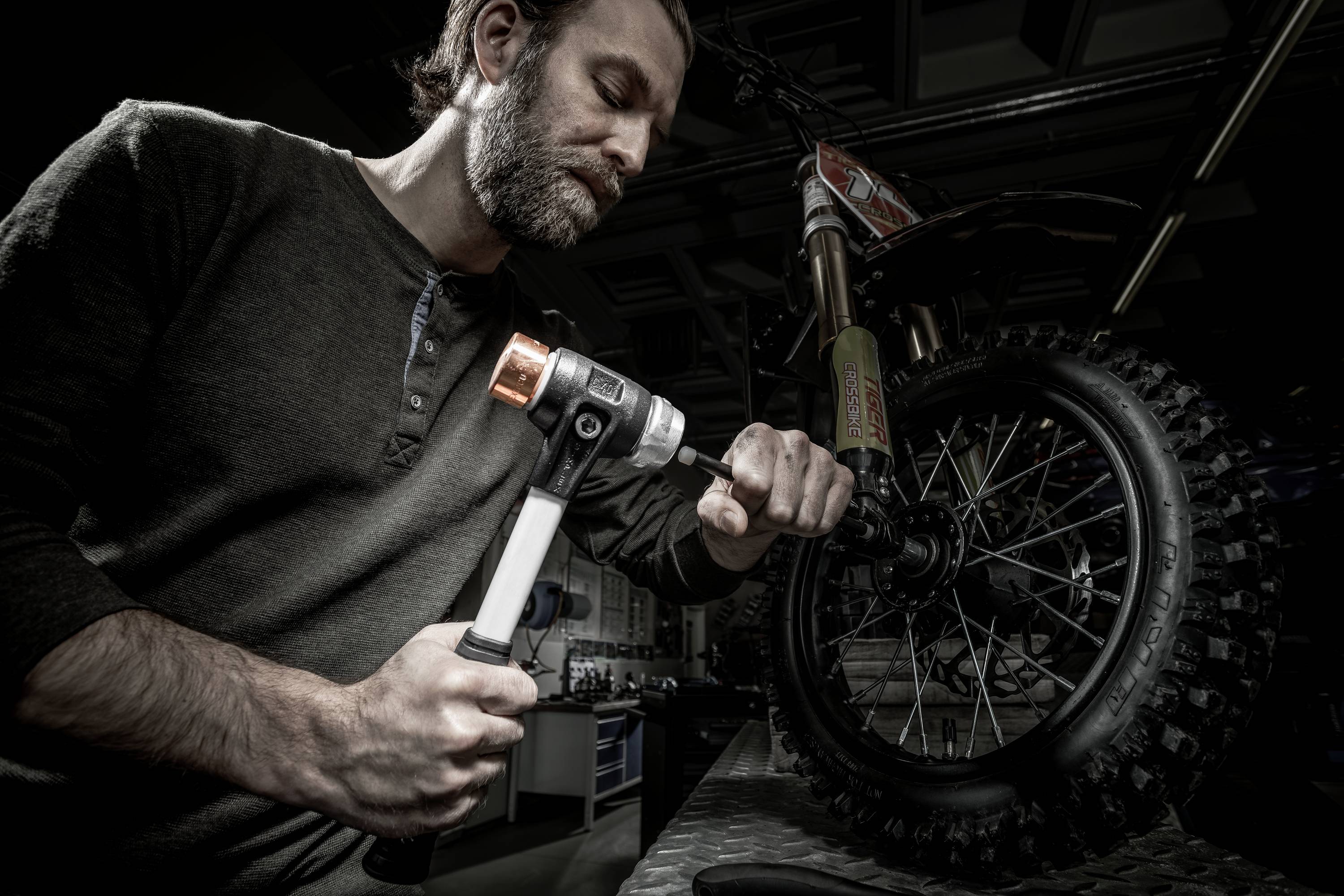 Muž opravuje motocykel kladivom v dielni. Dôraz je kladený na pneumatiku, pričom je kladivo zdvihnuté.