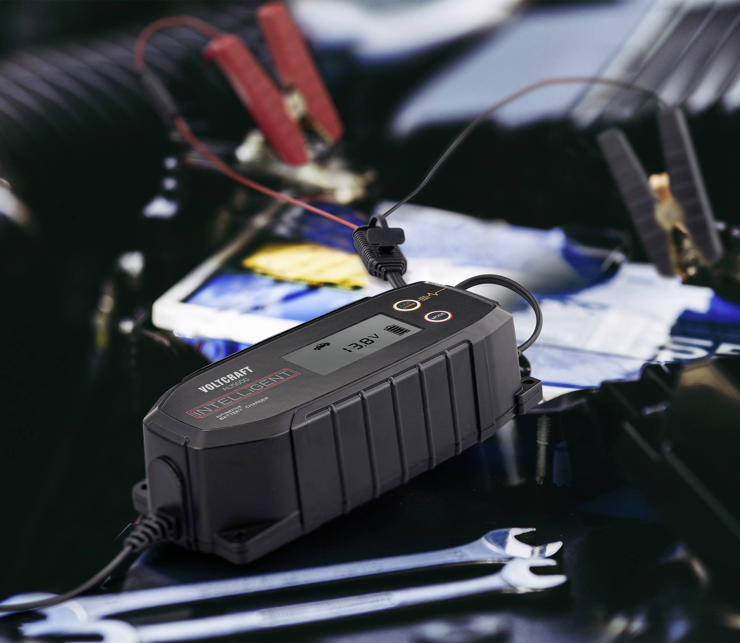 Nabíjačka batérie je pripojená ku svorkám autobatérie. Ukazuje napätie 13,6V. V pozadí je rozmazaný priestor motorového priestoru.
