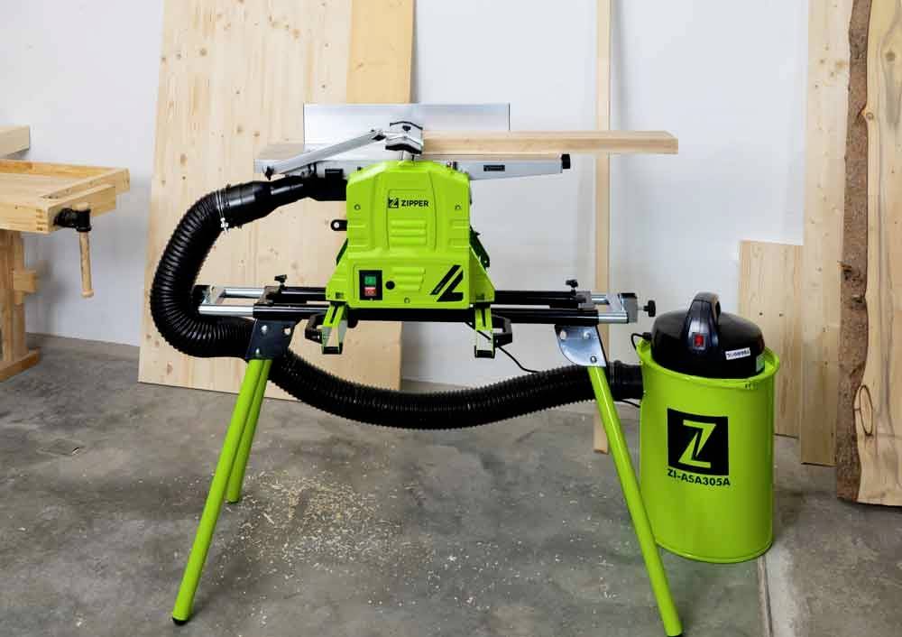 Zelená stolová okružná píla na stojane v dielni. Vedľa píly stojí zelený vysávač. V pozadí sú viditeľné drevené dosky.