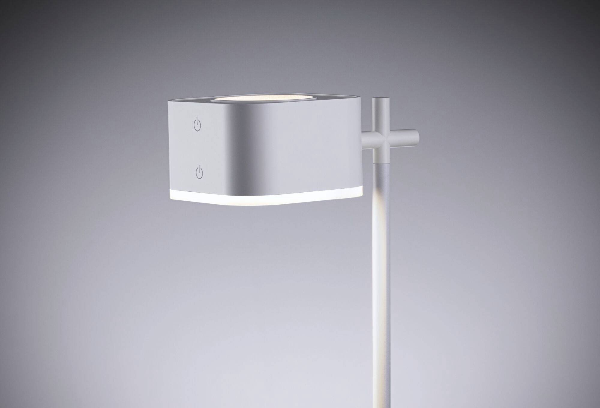 Strieborná moderná stolná lampa s minimalistickým dizajnom, zavesená na vertikálnej tyči, jemne žiari na sivom pozadí.
