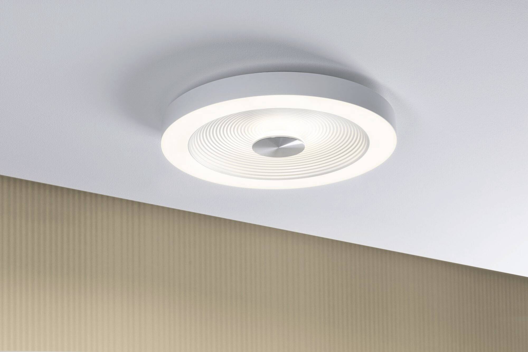 Moderné stropné svietidlo s bielym, kruhovým dizajnom jemne žiari zo stropu. Pozadie tvorí béžová stena.