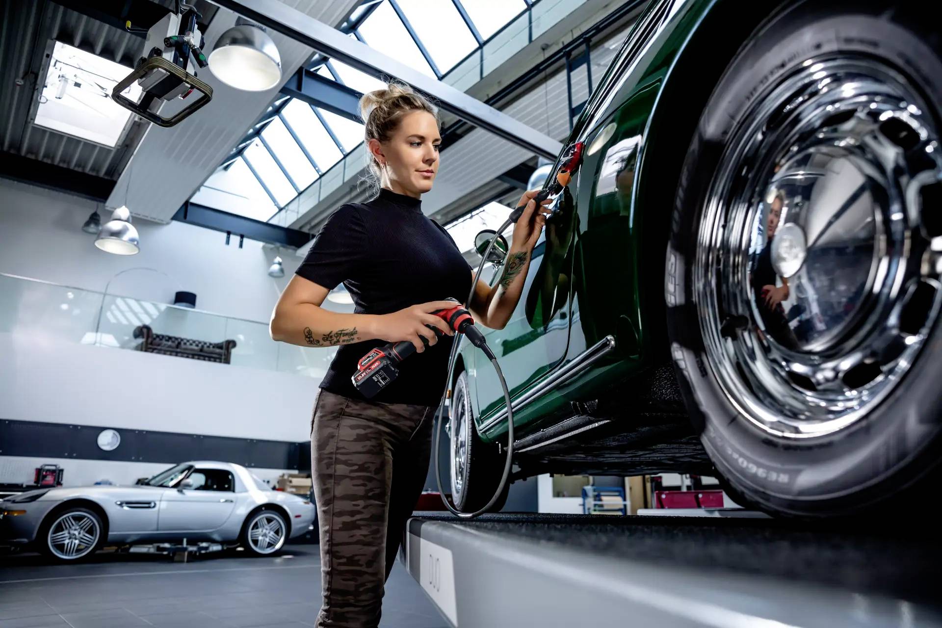 Žena leští tmavé auto v dielni; v pozadí stojí strieborné auto. Používa špeciálne leštiace náradie.