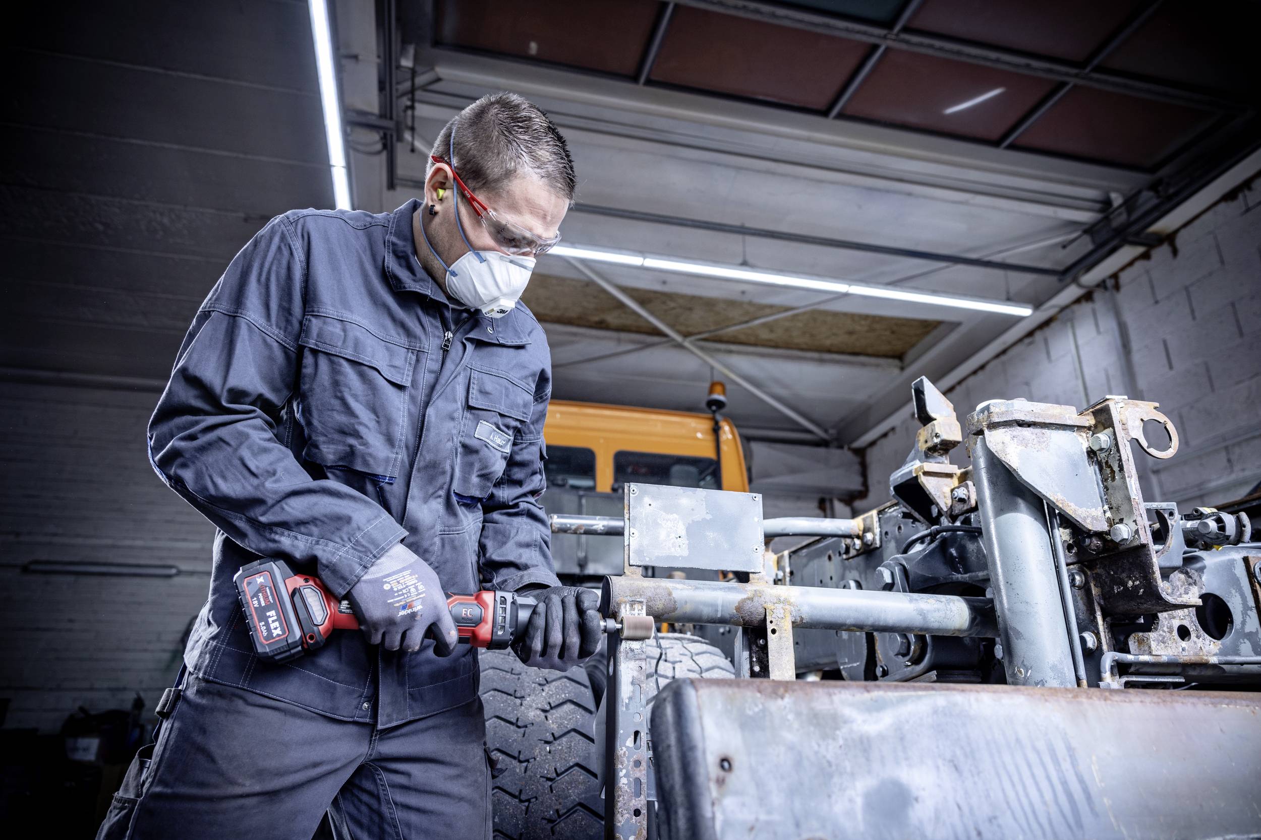 Muž v pracovnom oblečení, ochranných okuliaroch a mask pracuje s elektrickým náradím na vozidle v autoservise.