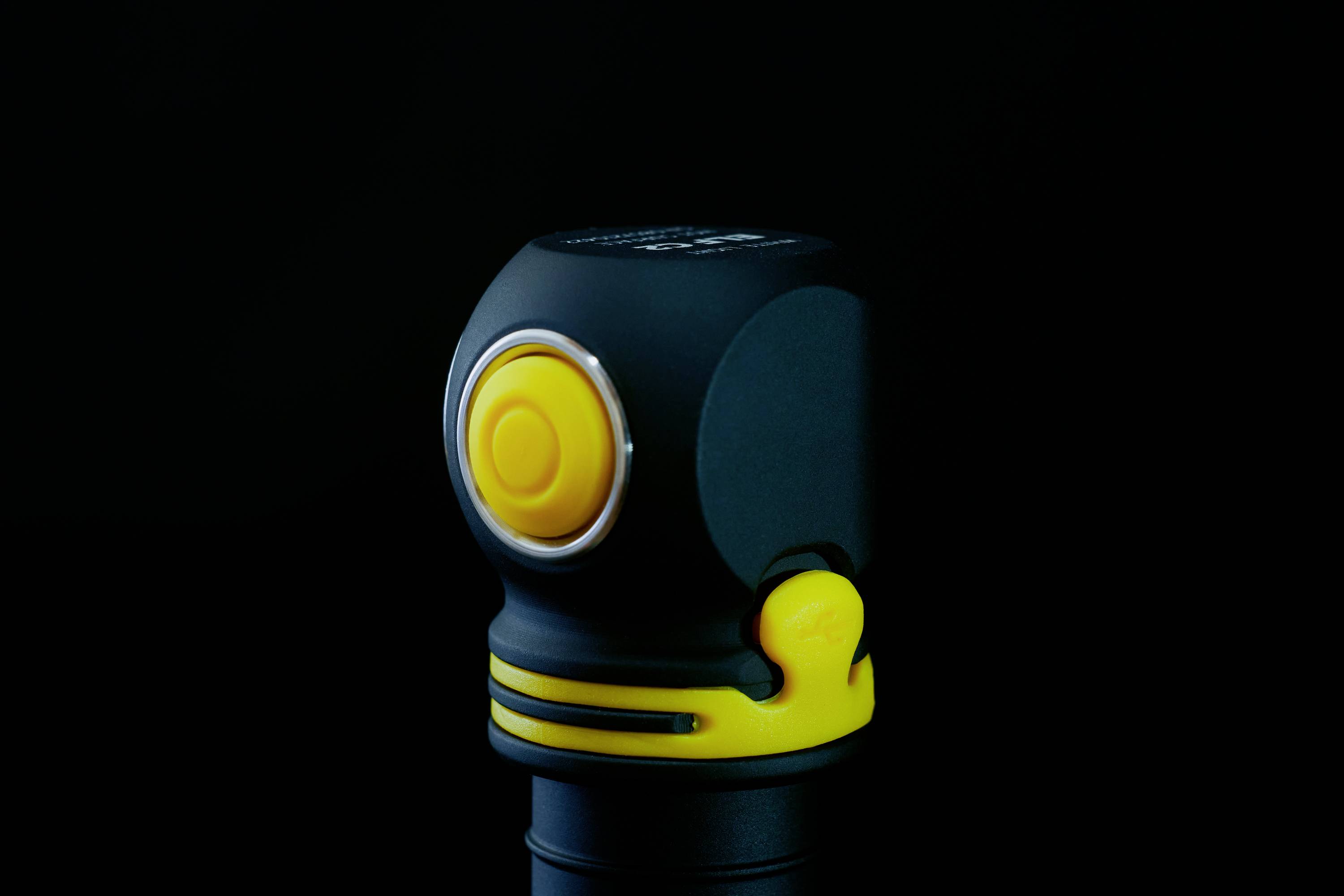 Čierna, kompaktná baterka so žltým spínačom na tmavom pozadí. Minimalistický dizajn.