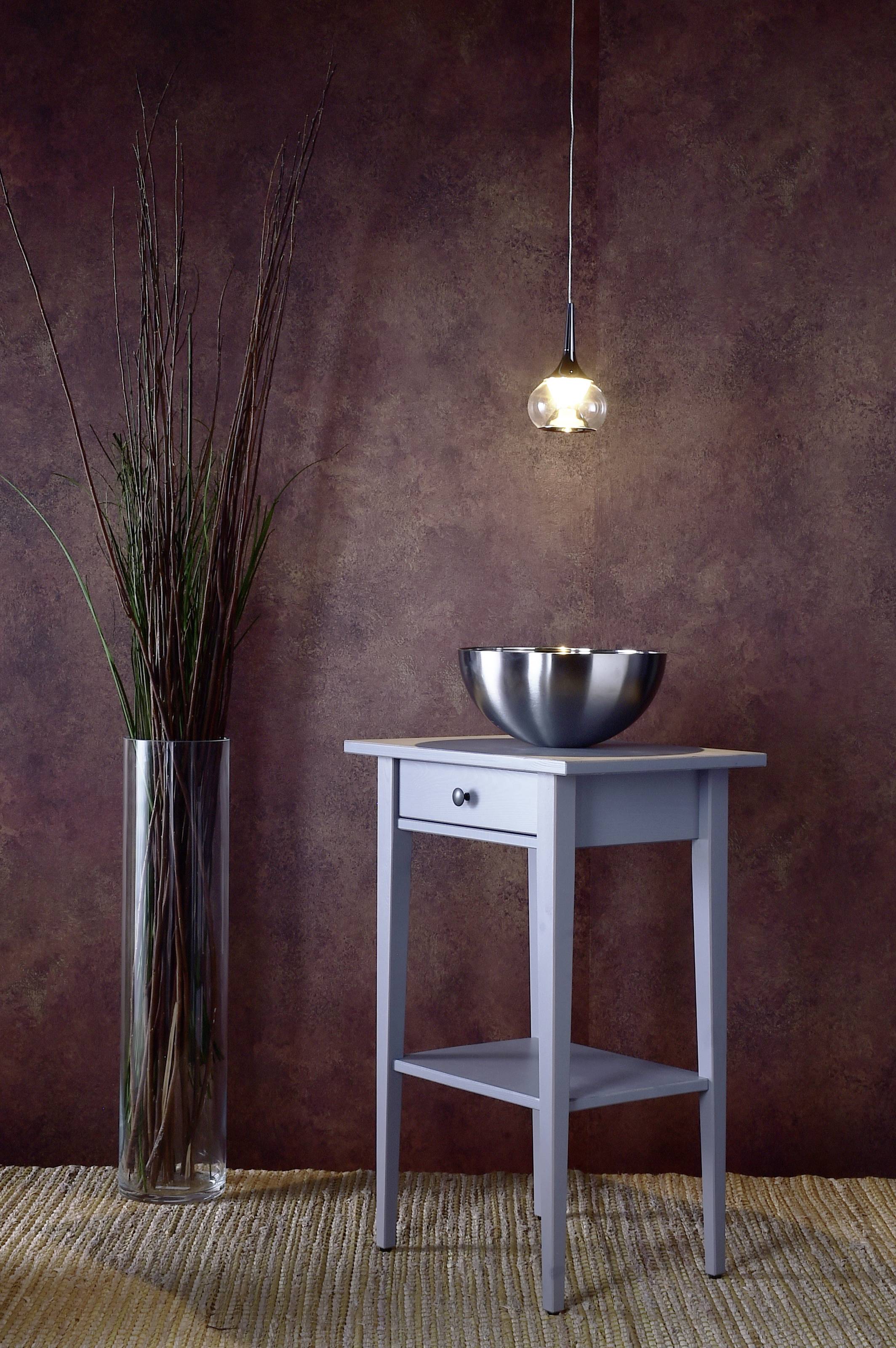 Stôl s misou, závesná lampa nad ním a vysoká váza s tenkými vetvami vedľa, stojí na koberci pred hnedou stenou.