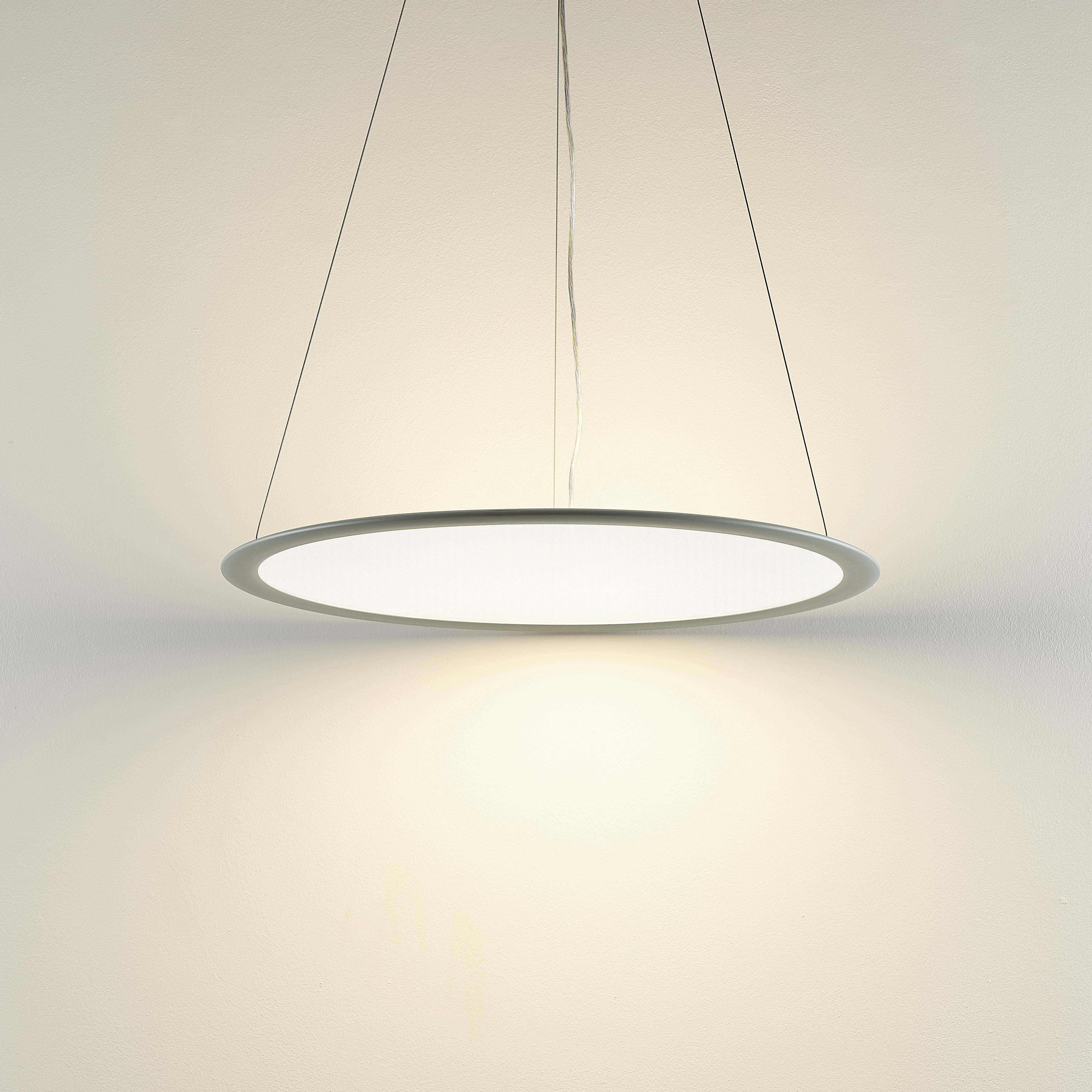 Okrúhla závesná lampa z kovu s rovnomerným rozptylom svetla, ktorá vyžaruje teplé biele svetlo na svetlú stenu.