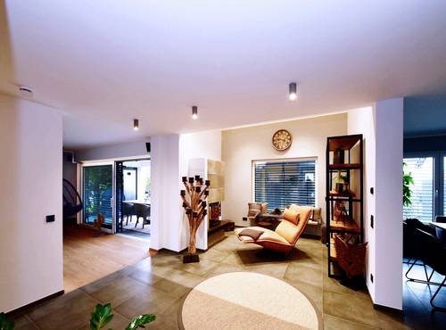 Moderná obývačka so svetlým osvetlením, pohodlným kreslom, hodinami na stene a sklenenými dverami vedúcimi do záhrady v pozadí.