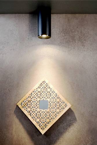 Lampa osvetľuje vzorový panel na stene. Lampa má valcovitý tvar a panel zobrazuje dekoratívny geometrický dizajn.