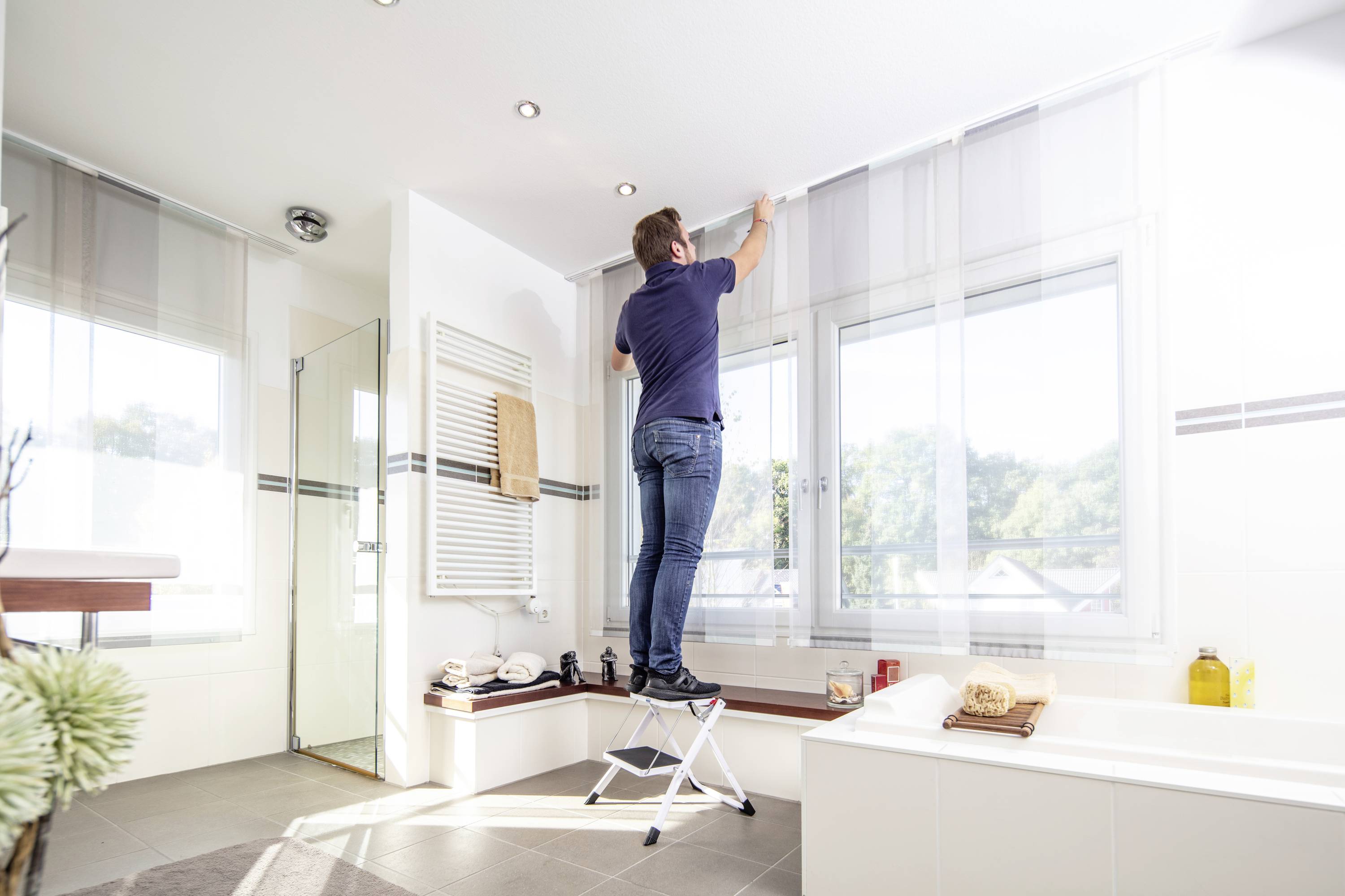 Osoba stojí na stoličke v kúpeľni a zavesuje alebo upevňuje záclony.