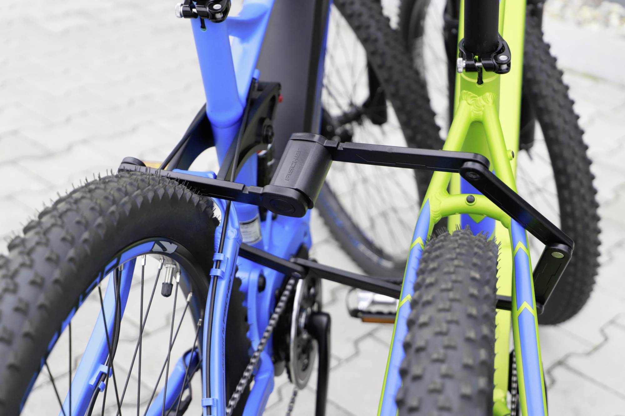 Dva bicykle sú zabezpečené čiernym skladacím zámkom na dlažobnej ceste. Bicykle majú hrubé, profilované pneumatiky.