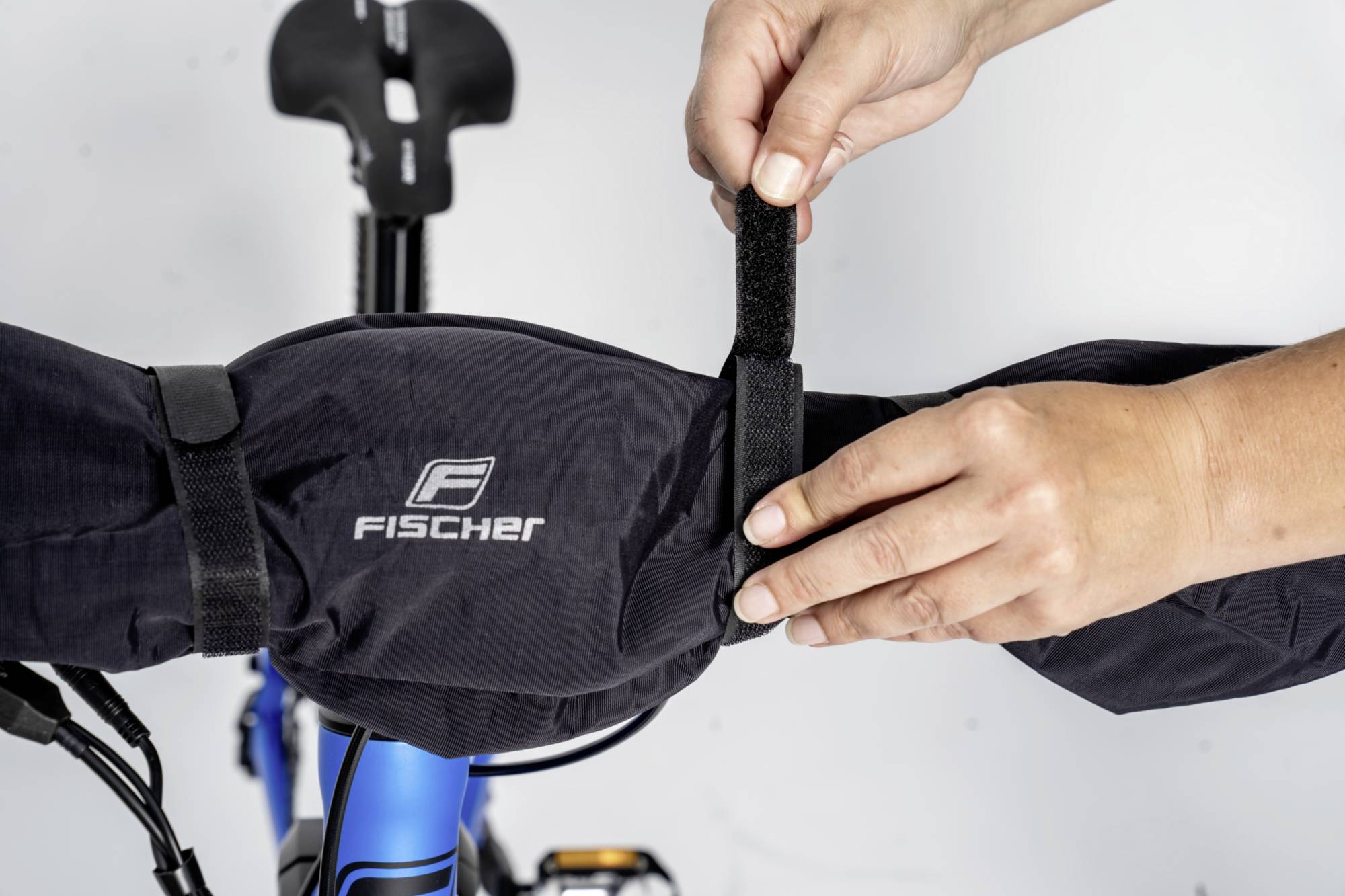 Osoba upevňuje cyklistickú tašku na riadidlá pomocou suchého zipsu. Taška nesie logo 'Fischer'. Riadidlá sú viditeľné v modrej farbe.