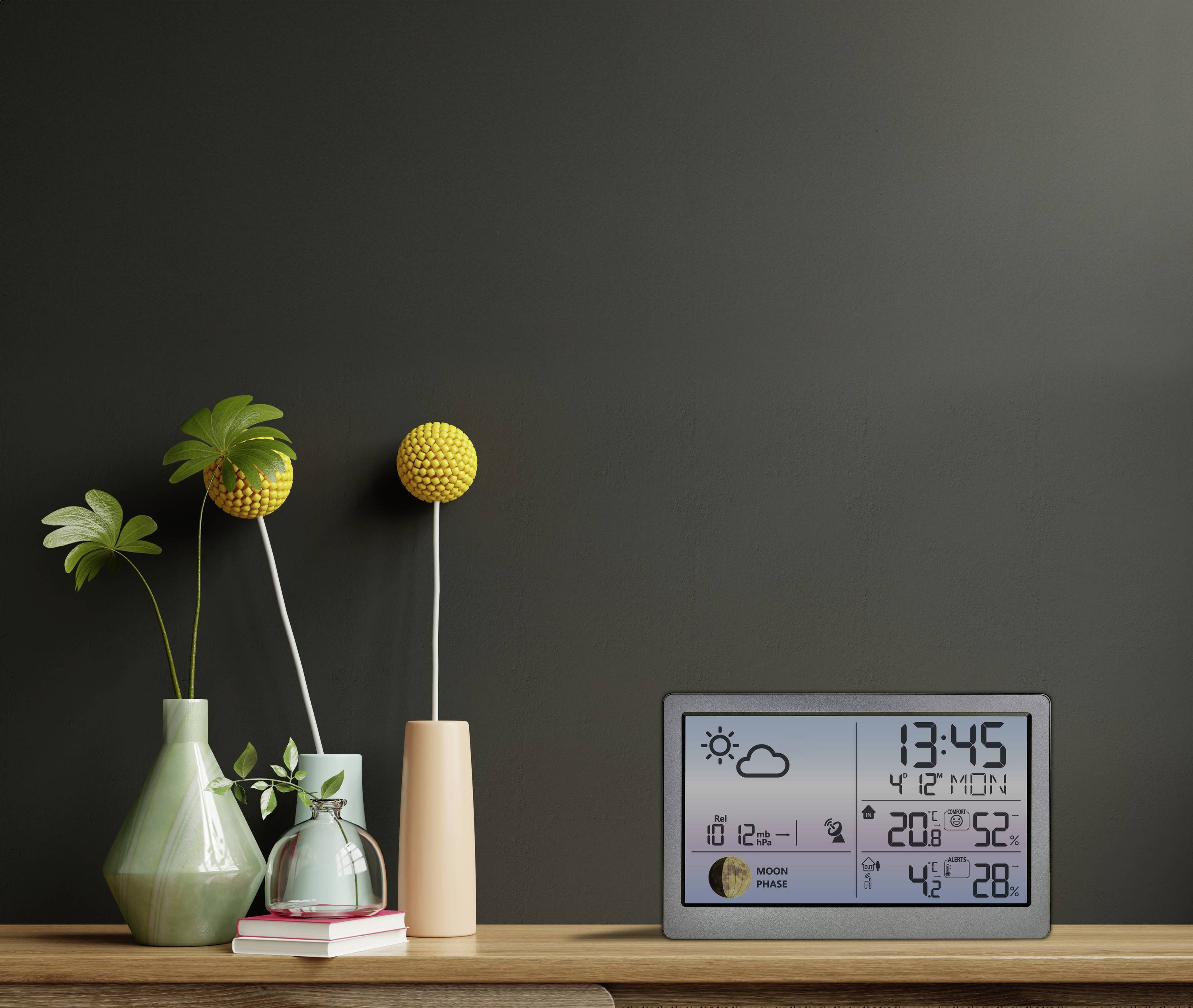Digitálny merač počasia a času na drevenom stole s zelenými a ružovými vázami. Čas: 13:45. Počasie: Slnečno, 20°C.