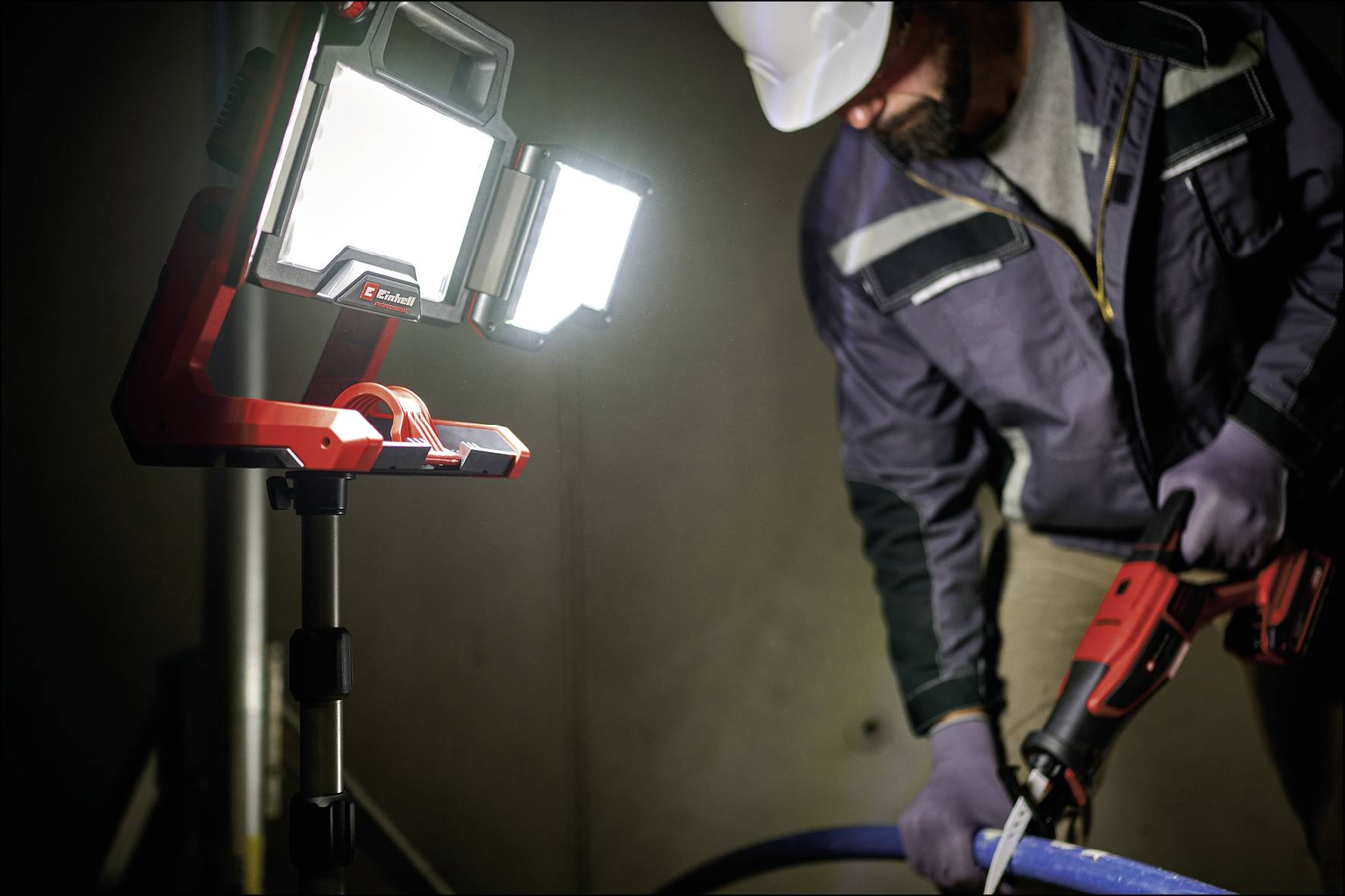 Robotník v helme a rukaviciach používa elektrický nástroj. Jasne svietiaca LED pracovná lampa osvetľuje scénu.